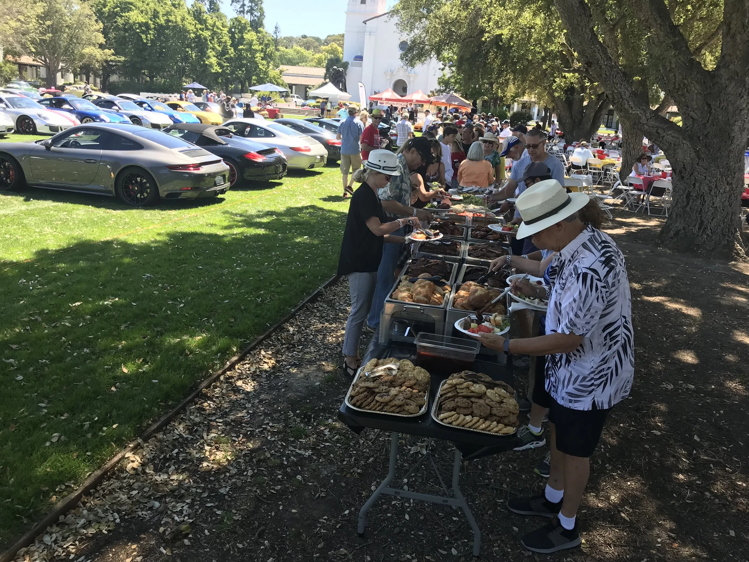 People standing around a table with catered food. Park and luxury cars in the background.