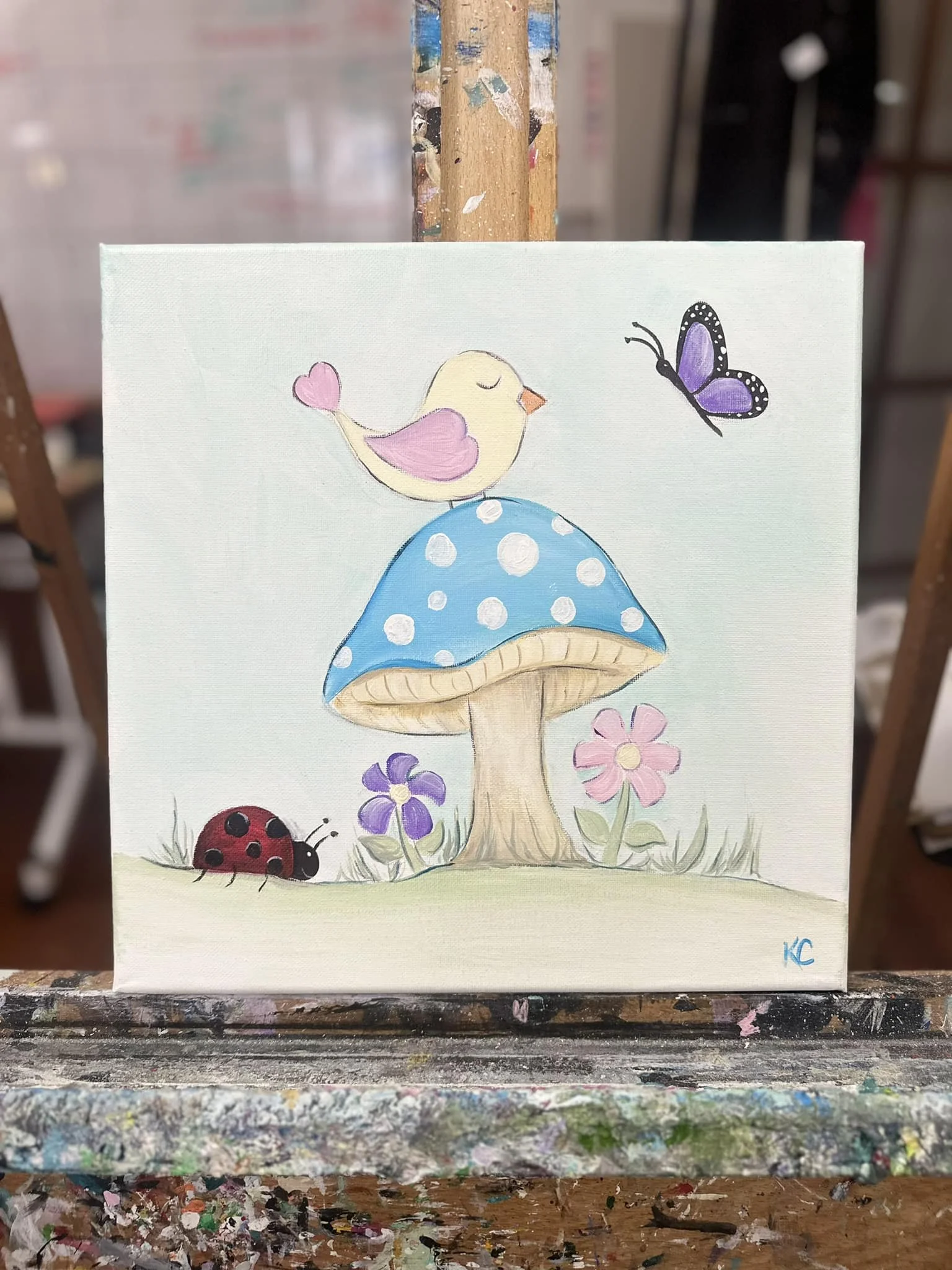 A painted canvas features a whimsical scene with a blue and white polka-dot mushroom at the center, surrounded by pink and purple flowers, a yellow bird with pink wings perched on top, a purple butterfly flying near the top right, and a ladybug on the grass near the bottom left.