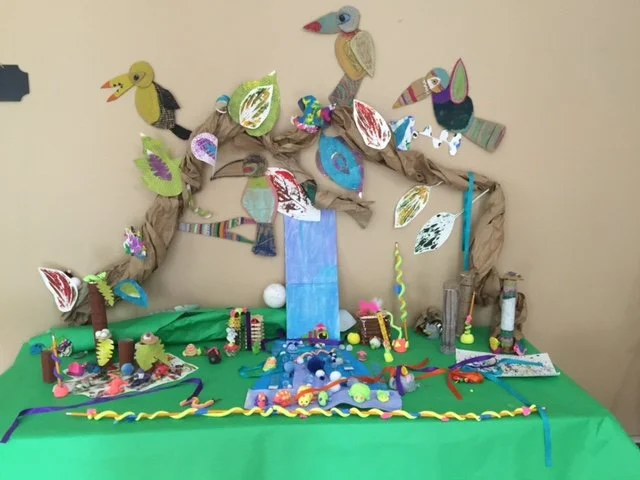 Colorful craft display with paper birds, leaves, a waterfall, and various small decorative items on a green tablecloth.