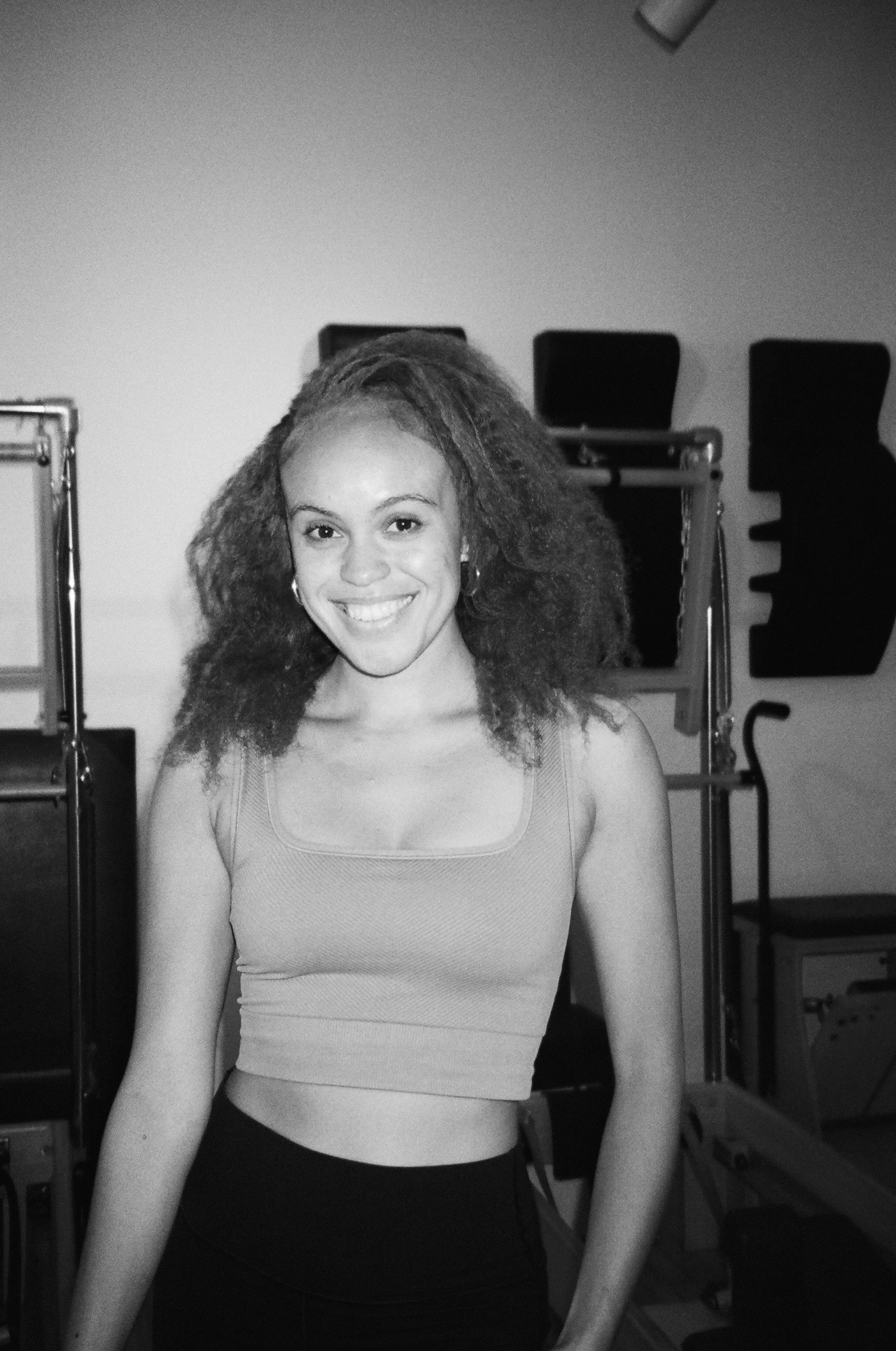 A smiling young woman with curly hair in a gym or physical therapy setting.