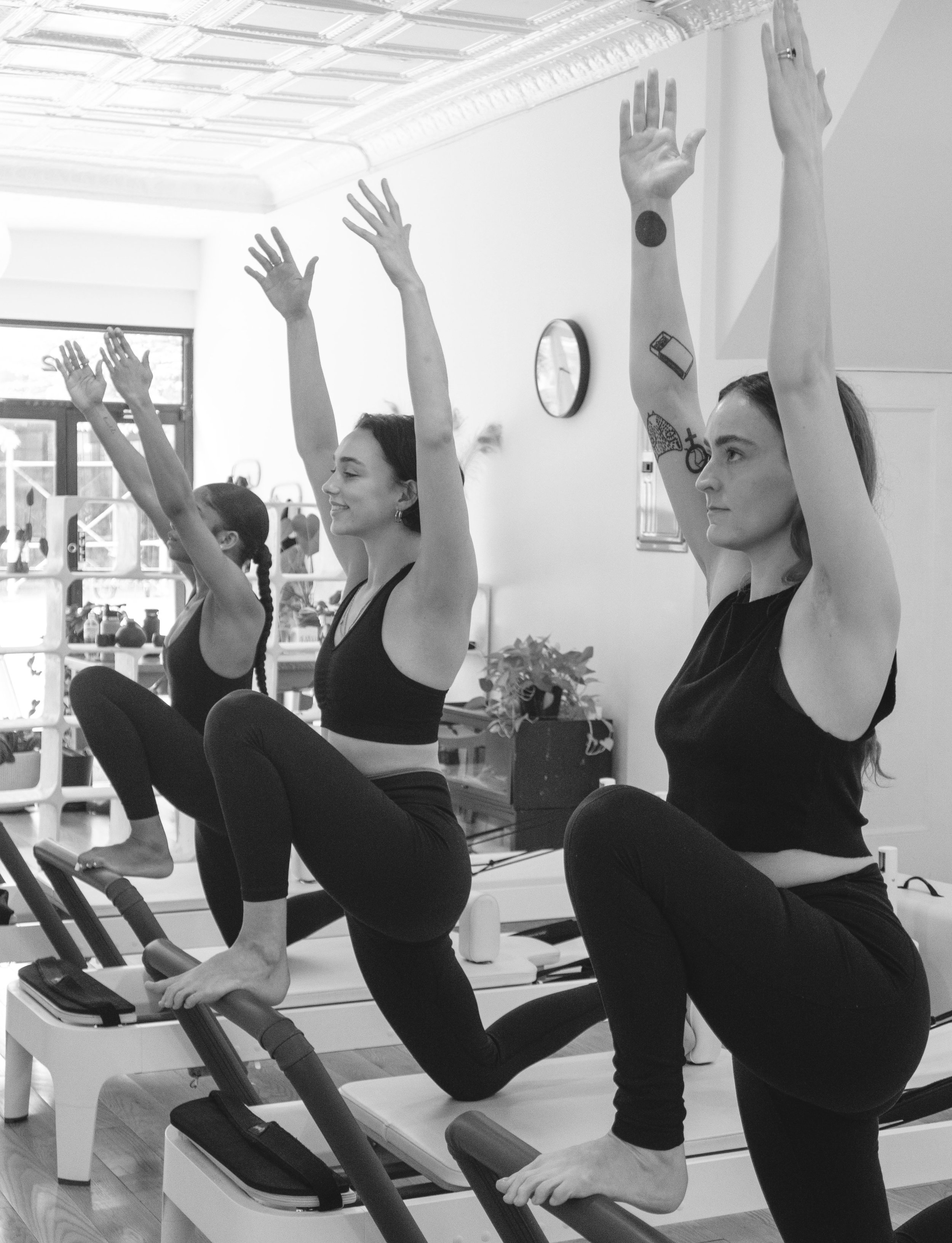 Three women in black workout clothes doing yoga on reformer machines in a fitness studio with large windows and a clock on the wall.