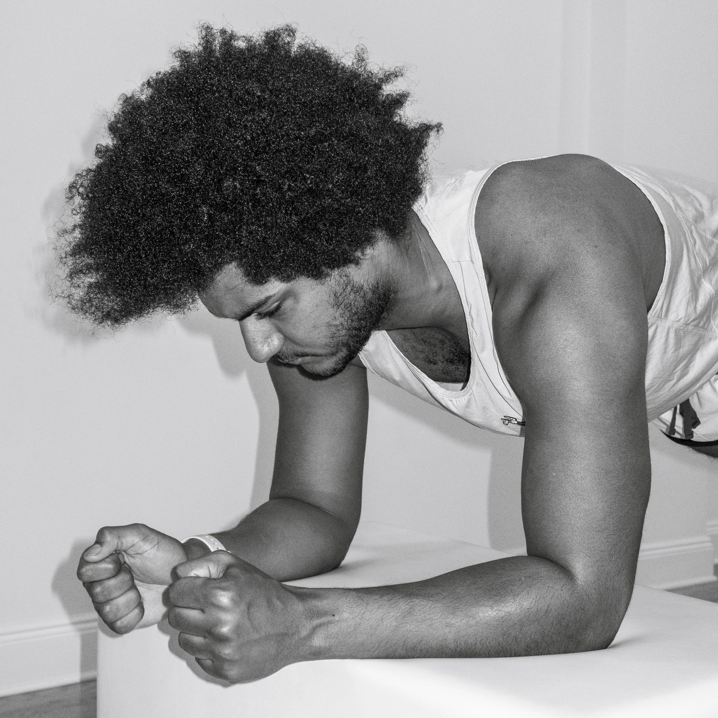 A man with curly hair and a beard is lying on a flat surface with his left arm bent, looking down and clenched in a fist, in a plank position.