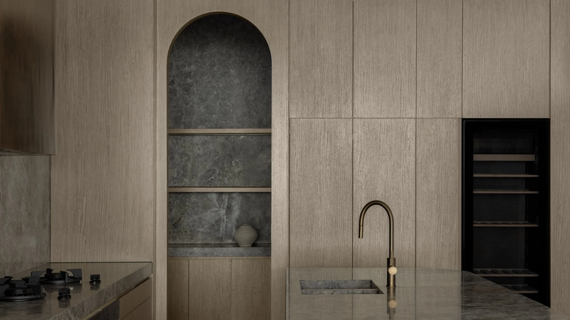Modern kitchen with beige cabinetry, a marble backsplash, a built-in wine cooler, a stone shelf with a vase, a gas stove, and a marble countertop with a gold faucet over a sink.