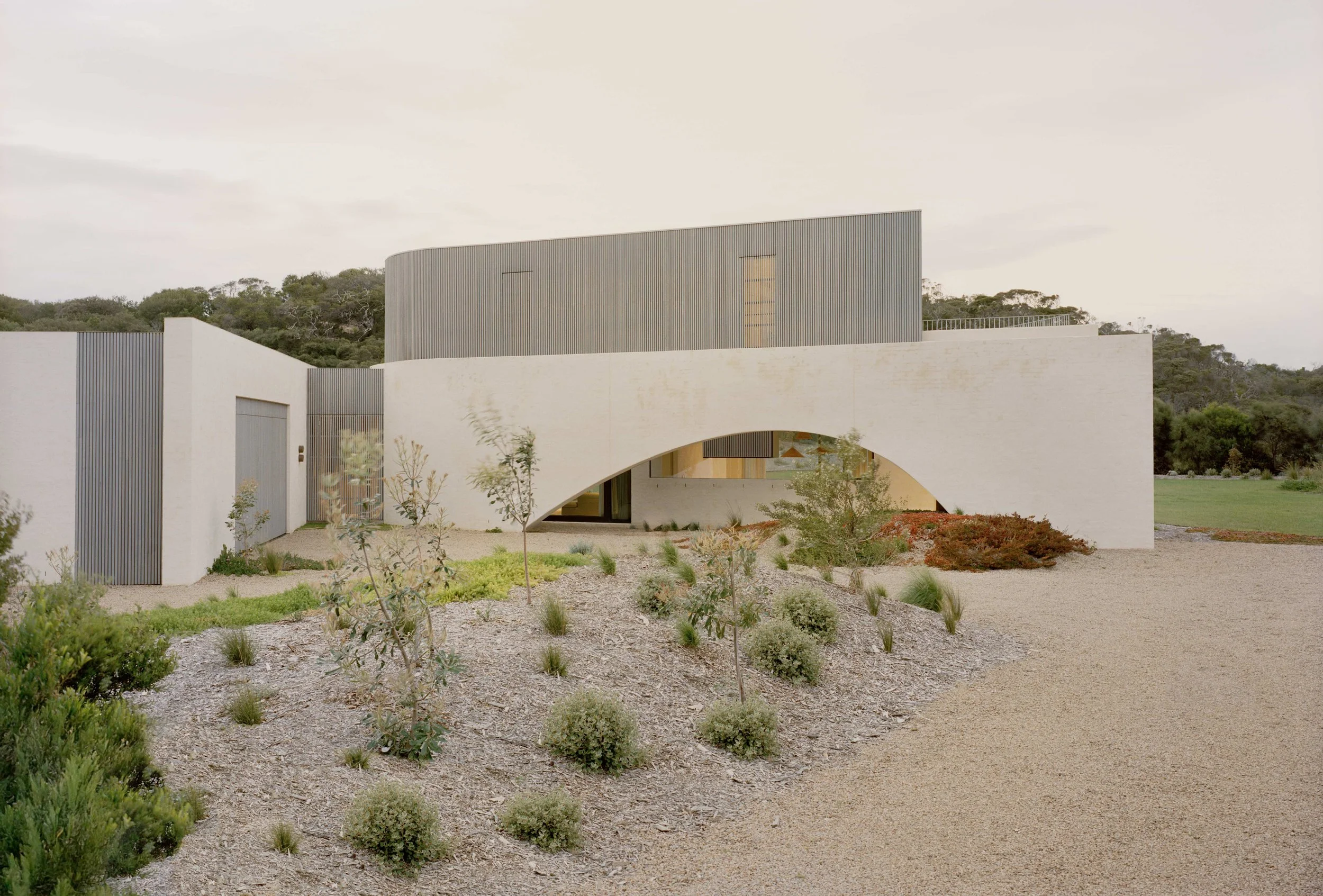 Modern house with white and gray exterior walls, arched entrance, surrounded by minimal landscaping with bushes, small trees, and gravel path, in a natural setting with hills in the background.