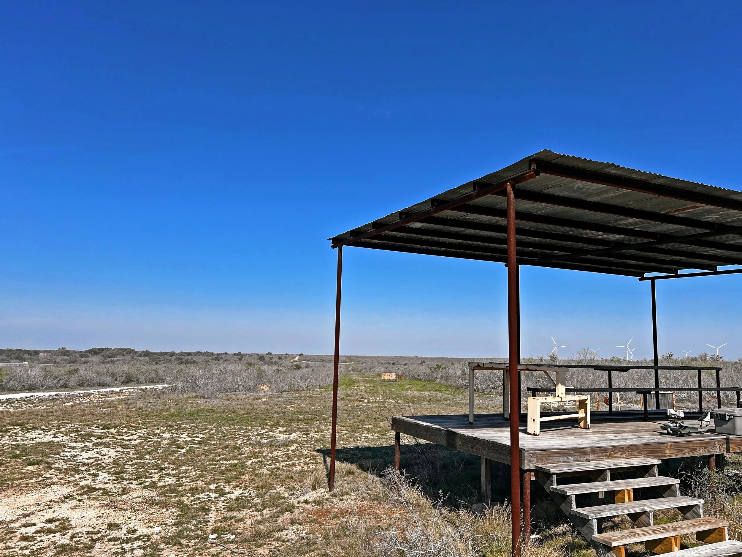 A small wooden structure with a corrugated metal roof on stilts in a vast, open, semi-arid landscape with wind turbines in the distance and a clear blue sky.