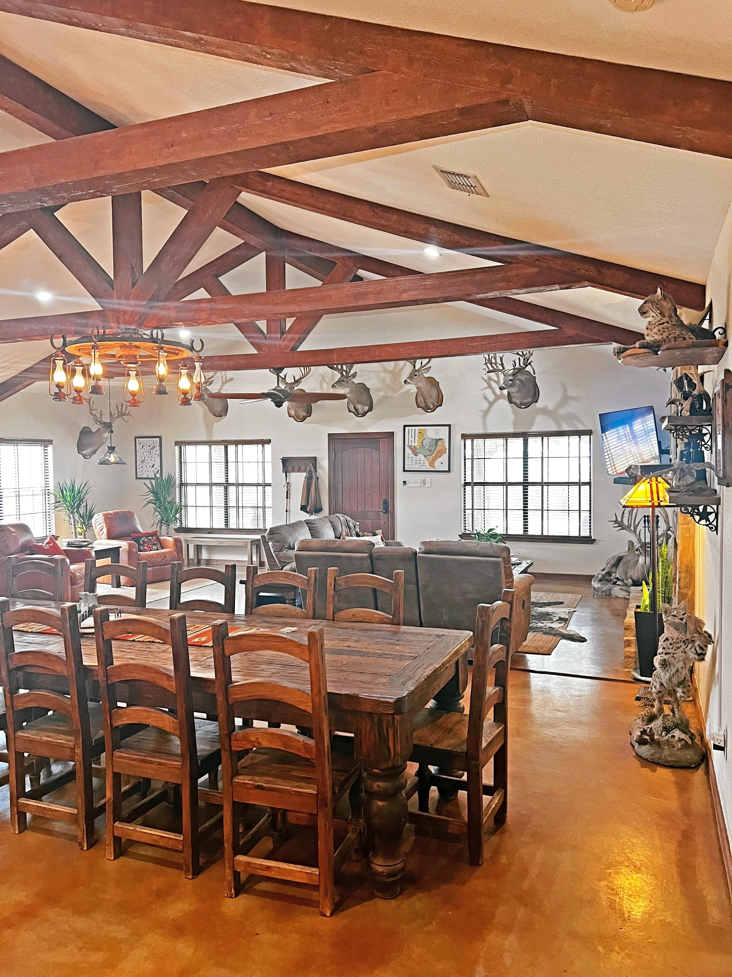 Living and dining room with deer and animal head decor, wooden beams on ceiling, large dining table, leather sofas, wall-mounted TV, and mountain lion figurines