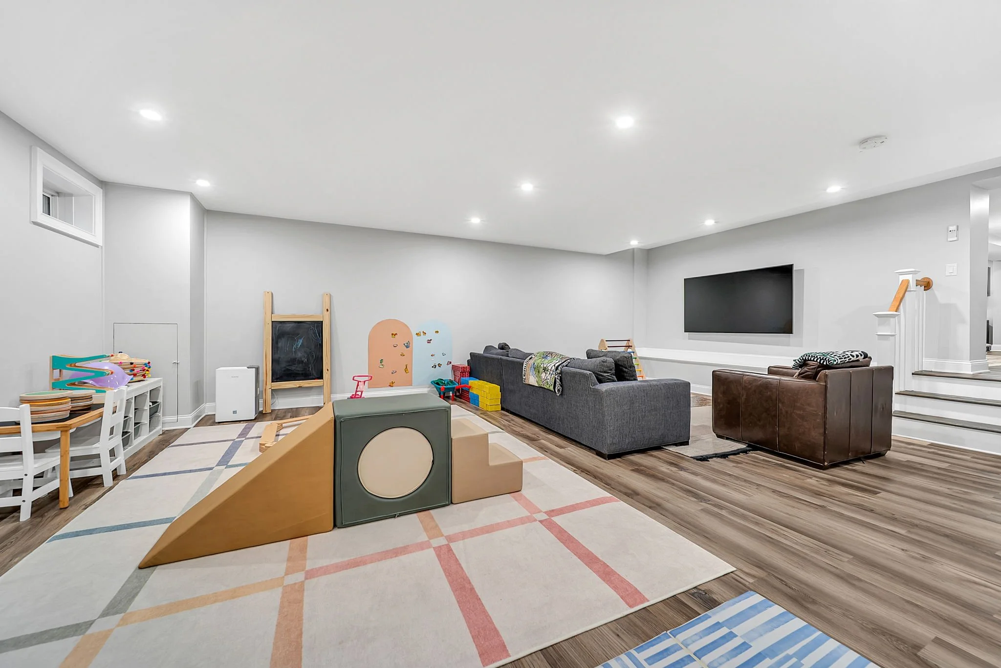 A spacious basement playroom with white walls, hardwood floors, and recessed lighting. It features children’s toys and climbing structures, seating area with sofas, a wall-mounted TV, and a staircase leading upstairs.