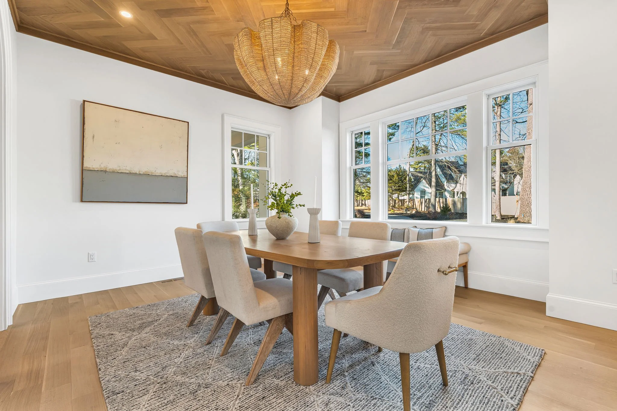 A dining room with a wooden table, six beige upholstered chairs, a textured area rug, large windows, a ceiling light, and wall art.