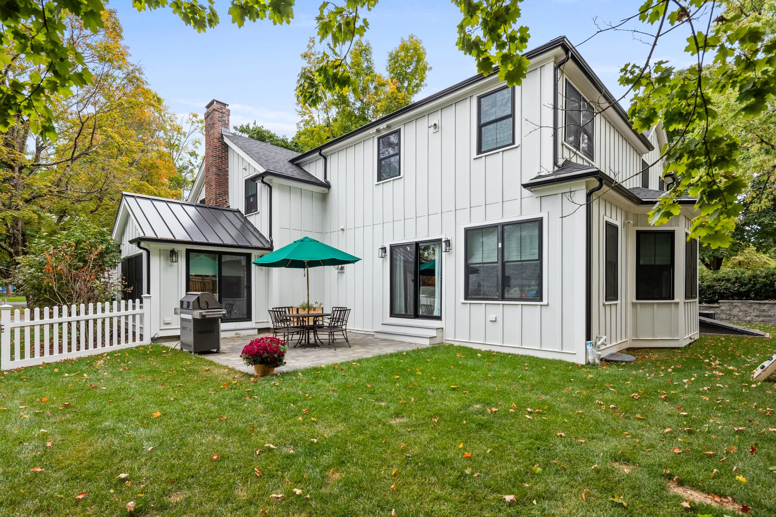 Backyard view of a two-story white house with black window frames, a patio with a table, four chairs, and a green umbrella, surrounded by green grass and trees with autumn foliage.