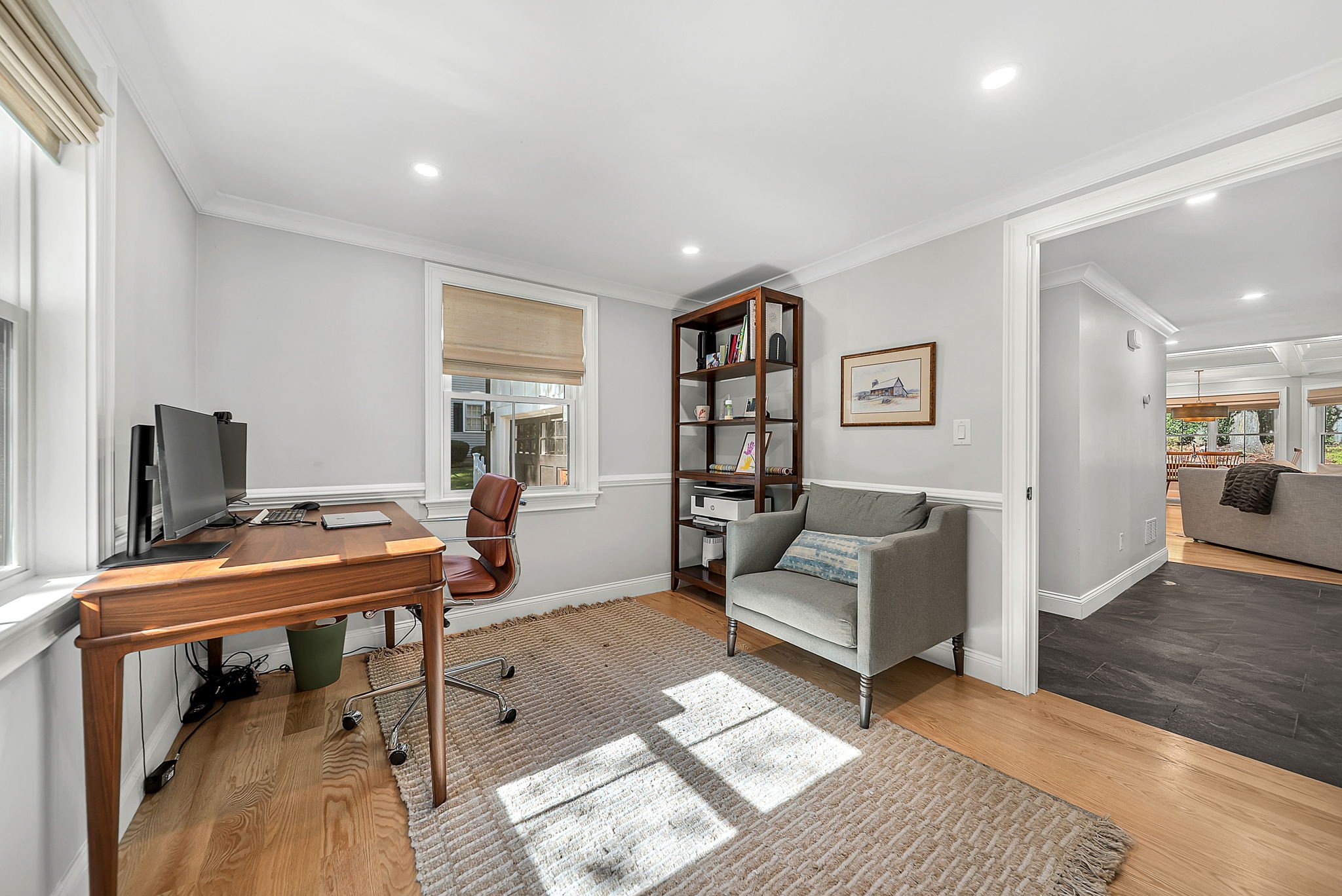 Home office with a wooden desk, computer, and office chair near a window; gray armchair, bookshelf, and framed art on the wall; sunlight on hardwood floor and a rug.