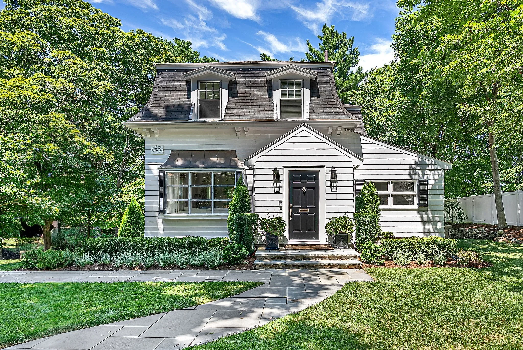 Front view of a two-story house with white siding, black front door, and black window shutters. The house has a curved staircase leading to the entrance, landscaped front yard with green grass, shrubs, and trees. Blue sky with some clouds visible in the background.