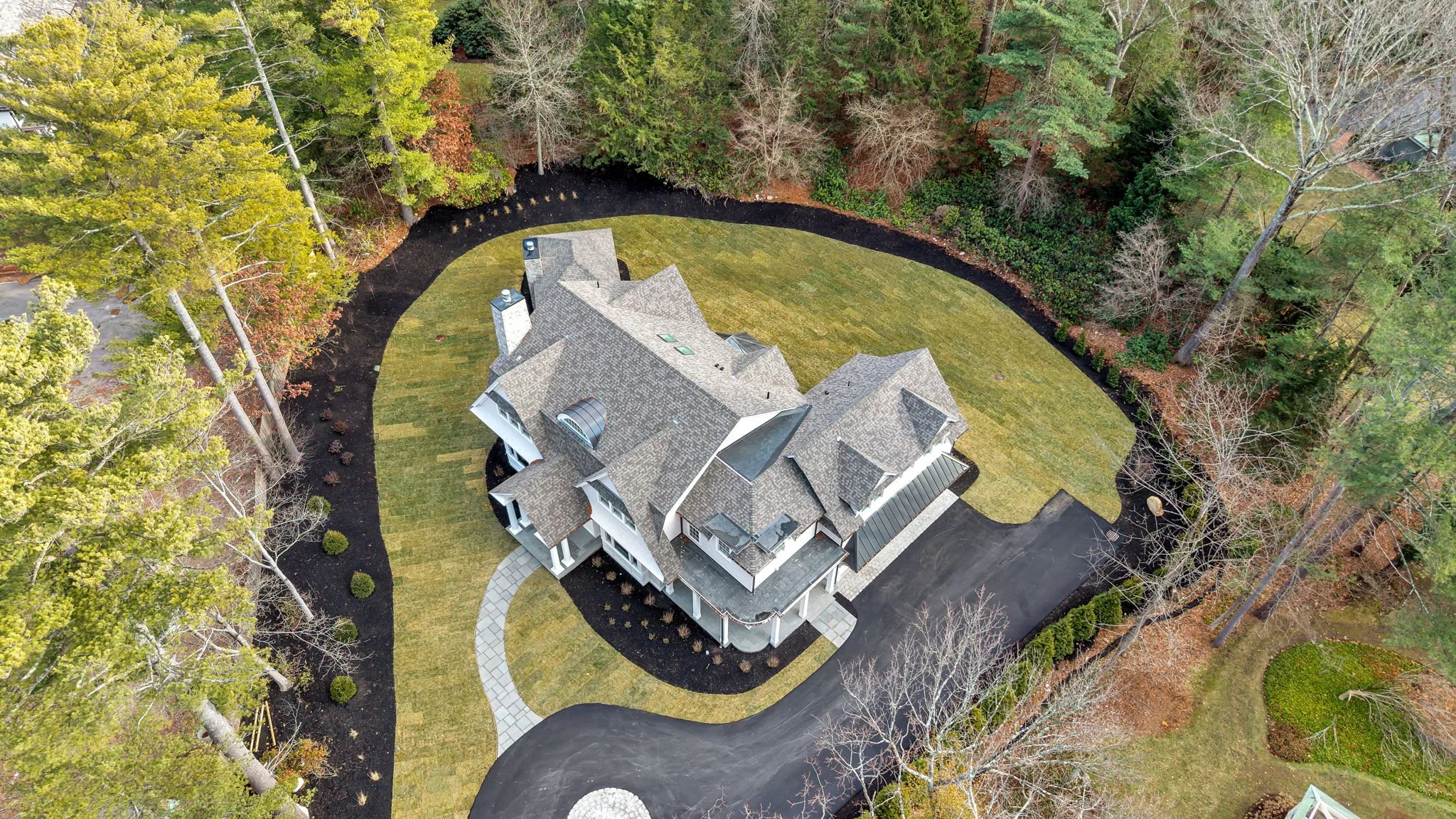 An aerial view of a large, multi-story house with a gray shingled roof, surrounded by a curved driveway, a grassy lawn, and tall trees in various seasons of leaf change.