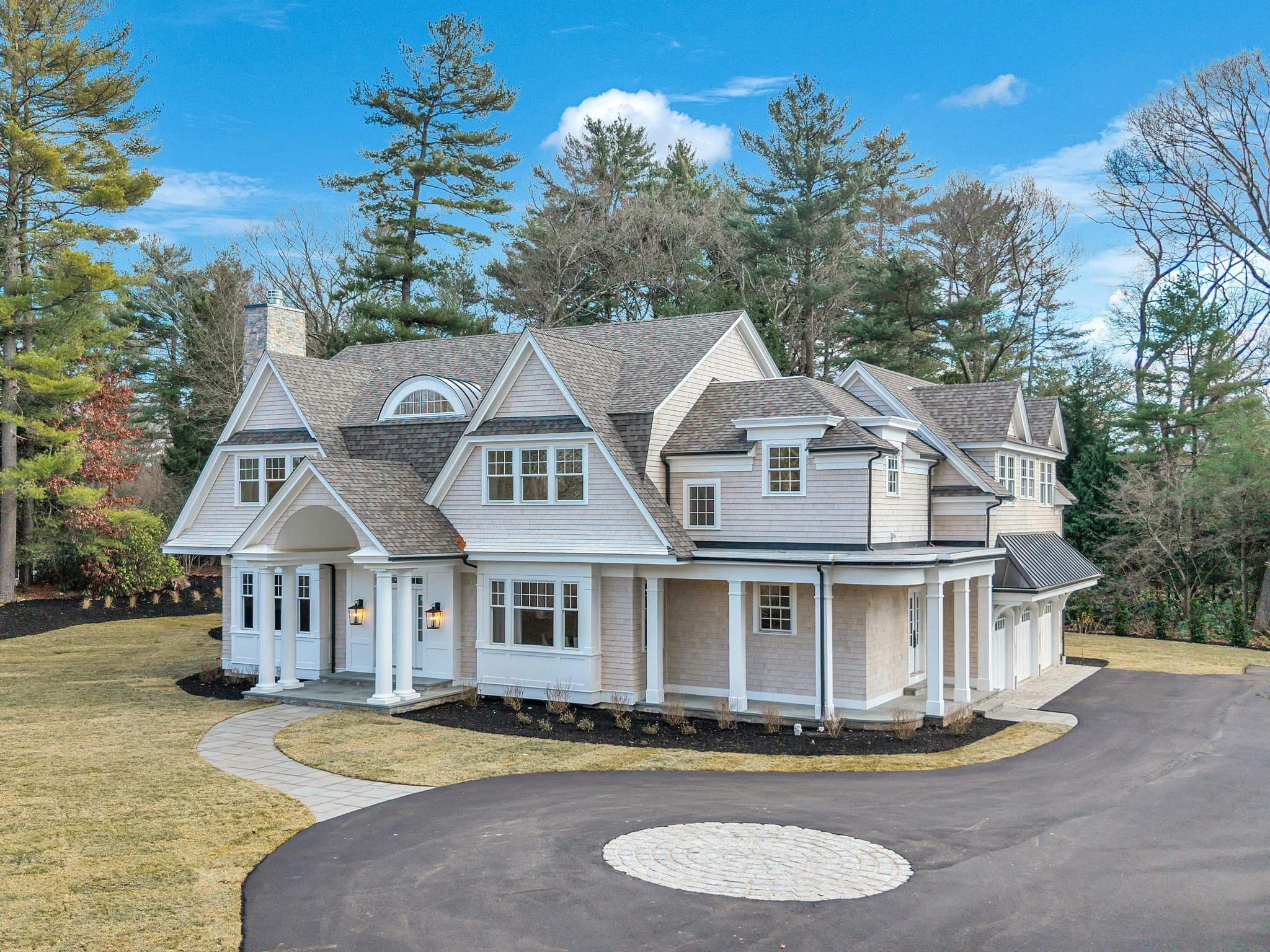Large white suburban house with multiple gables, porch with columns, surrounded by trees and a paved driveway.