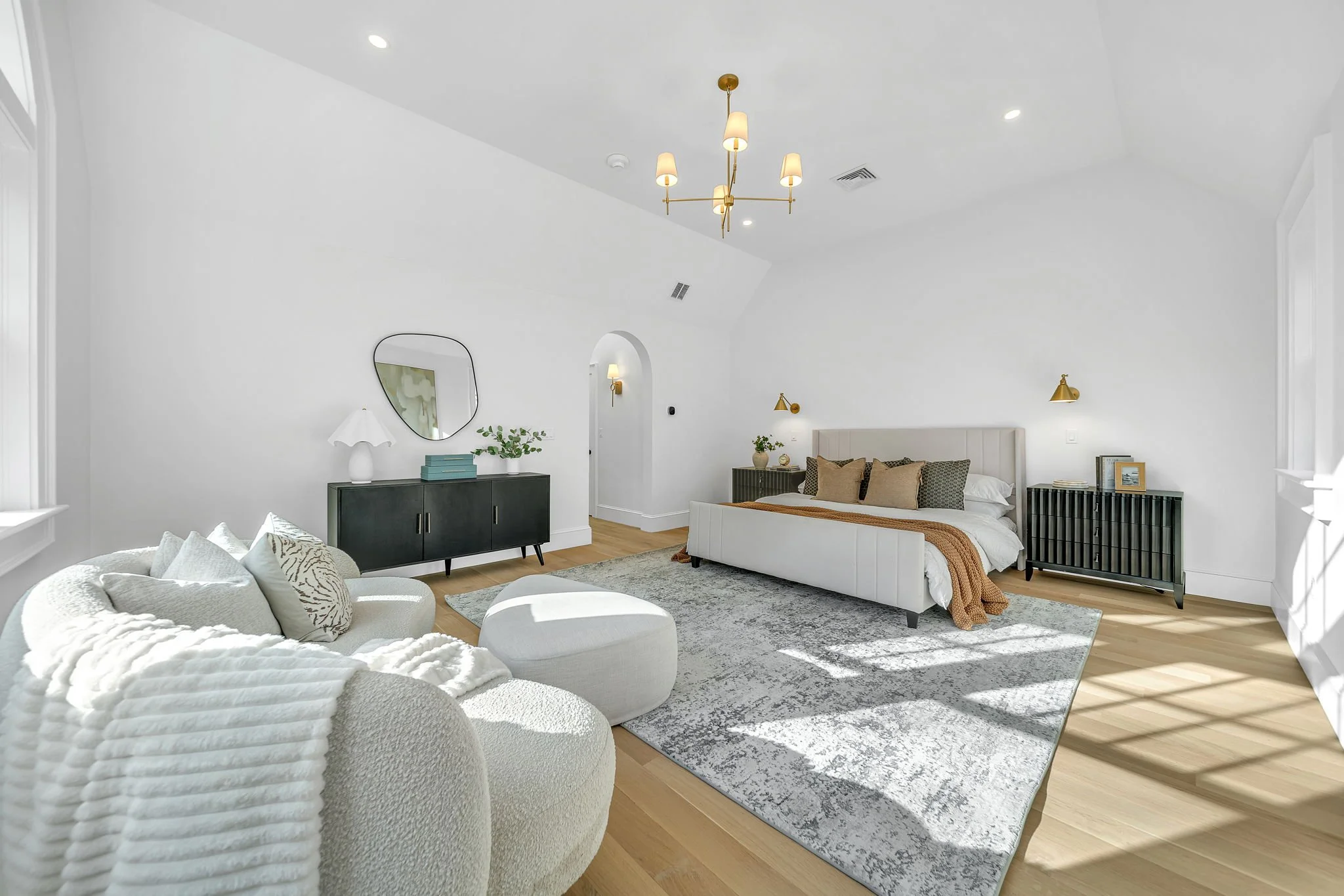 Bright, modern bedroom with white walls, light wood flooring, a bed with beige and brown pillows, black side tables with gold wall sconces, a white sofa with textured cushions, and a black sideboard with a mirror, lamp, books, and a plant.