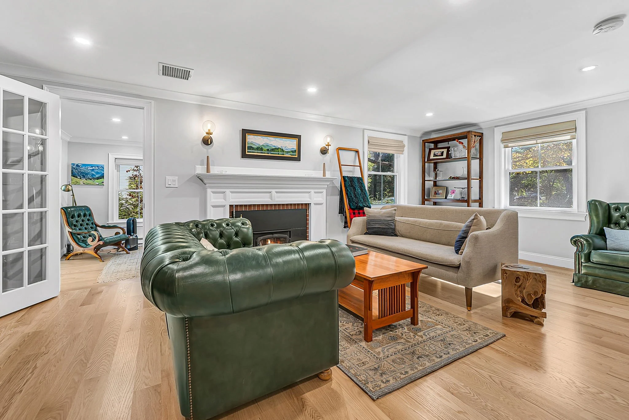Living room with white walls, hardwood floors, a fireplace, and vintage green leather sofas. There are multiple windows, a wooden bookshelf, and various decorative items.