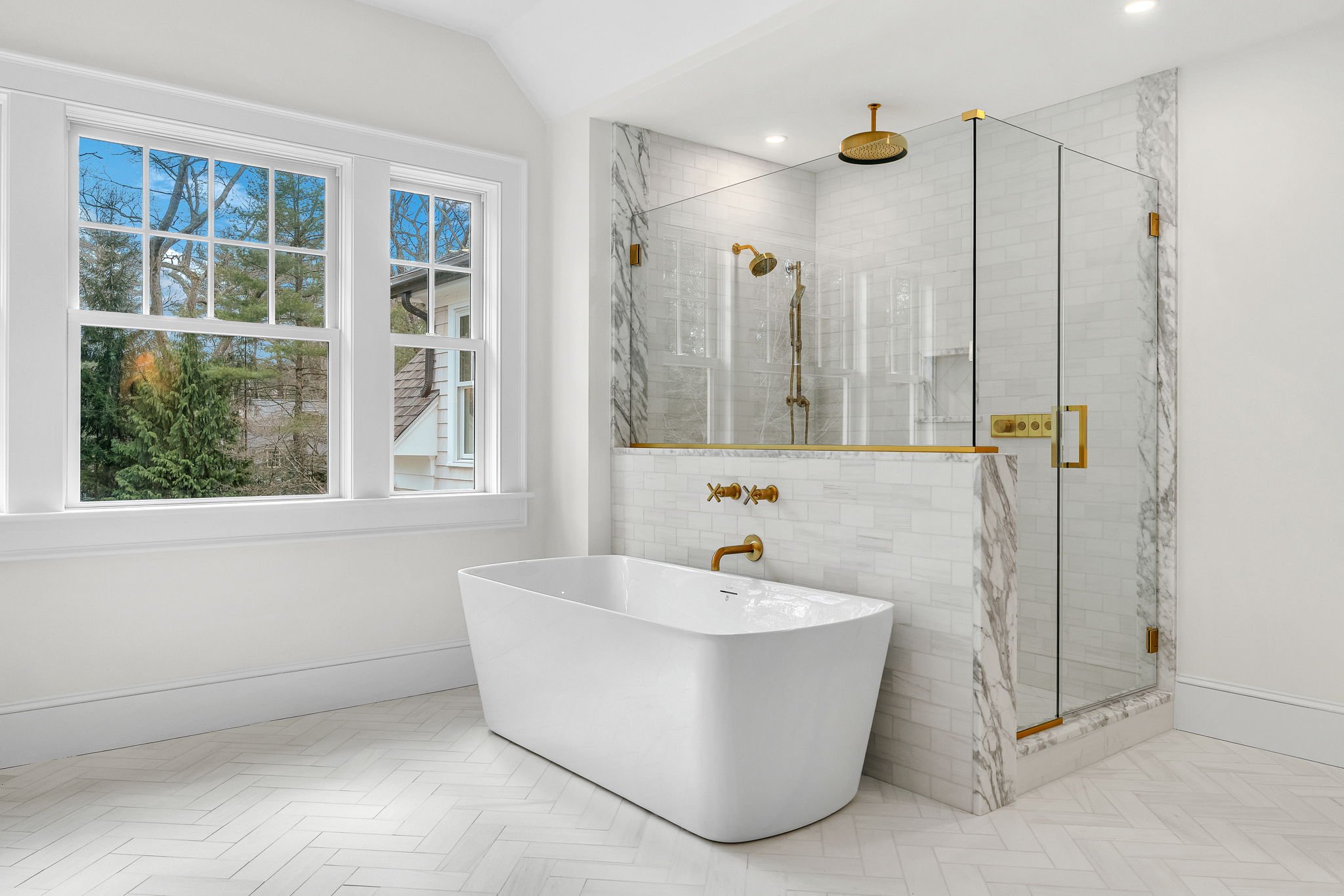 Modern bathroom with white freestanding bathtub, large window, and glass-enclosed shower with gold fixtures, marble accents, and white subway tile walls.