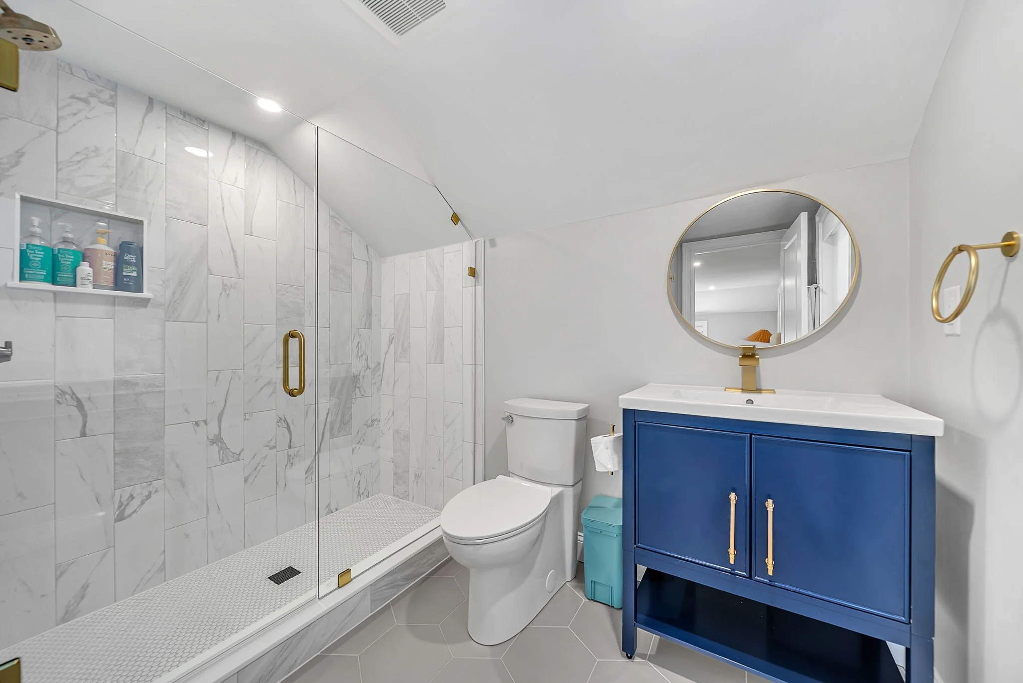 Modern bathroom with a walk-in shower with marble tiles, a white toilet, a blue vanity with gold handles, and a circular mirror with a gold frame on white walls.
