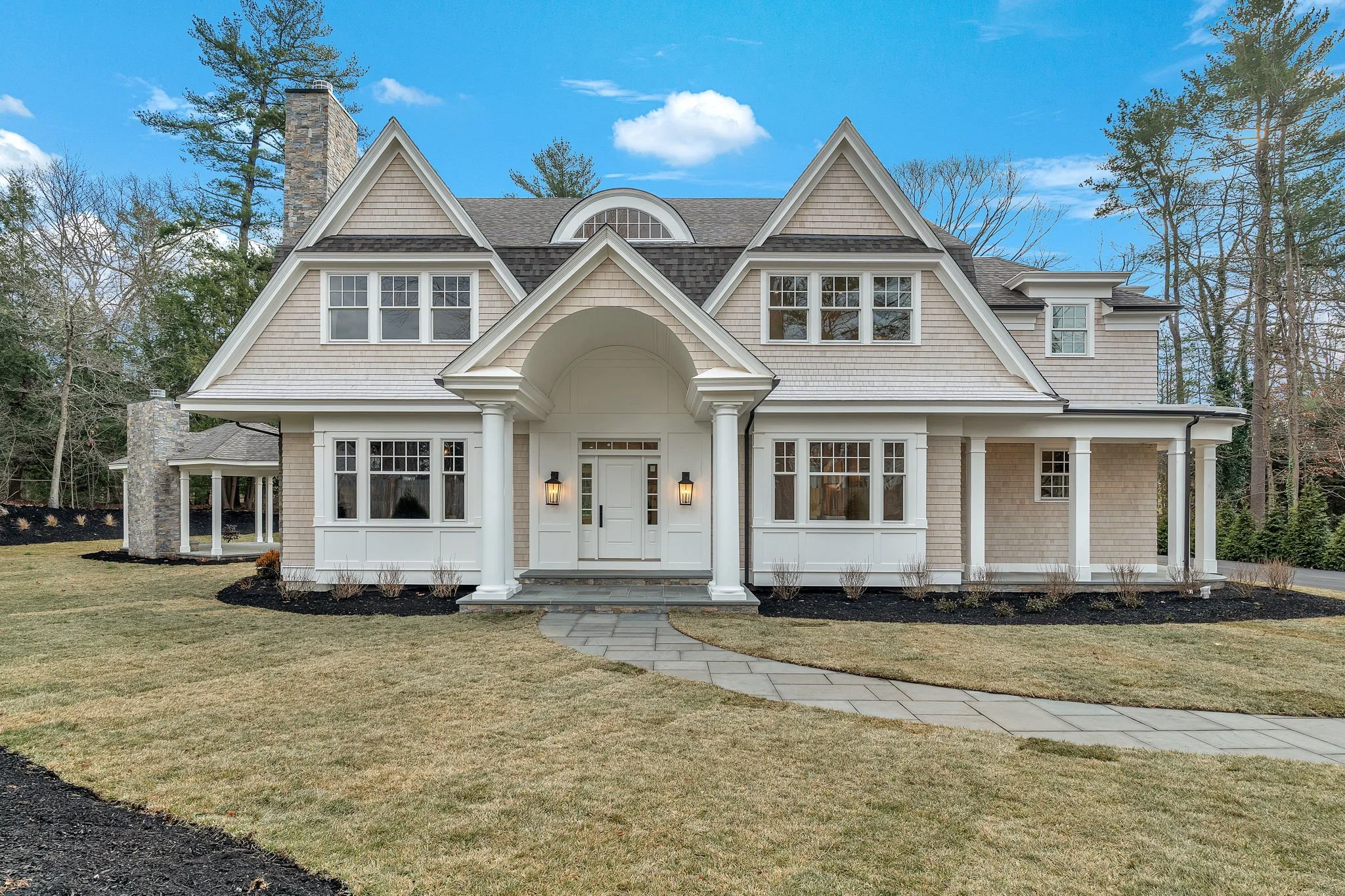 Front view of a large, two-story house with beige shingles, white trim, and a gray shingle roof, surrounded by a lawn and trees, under a blue sky with clouds.