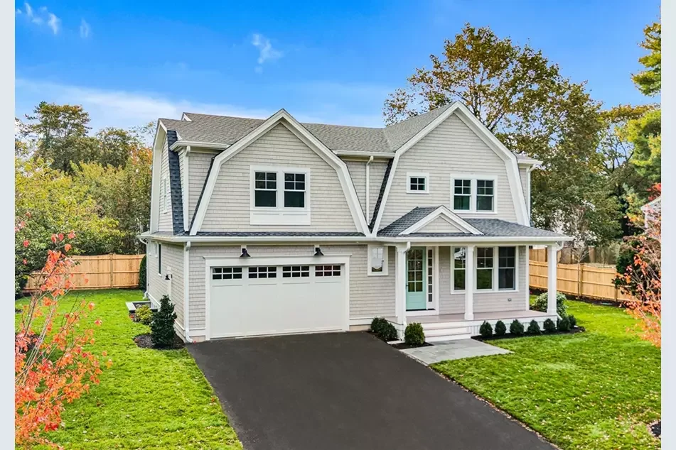 A two-story suburban house with white siding, gray roof, and a front porch, surrounded by a green lawn and trees.