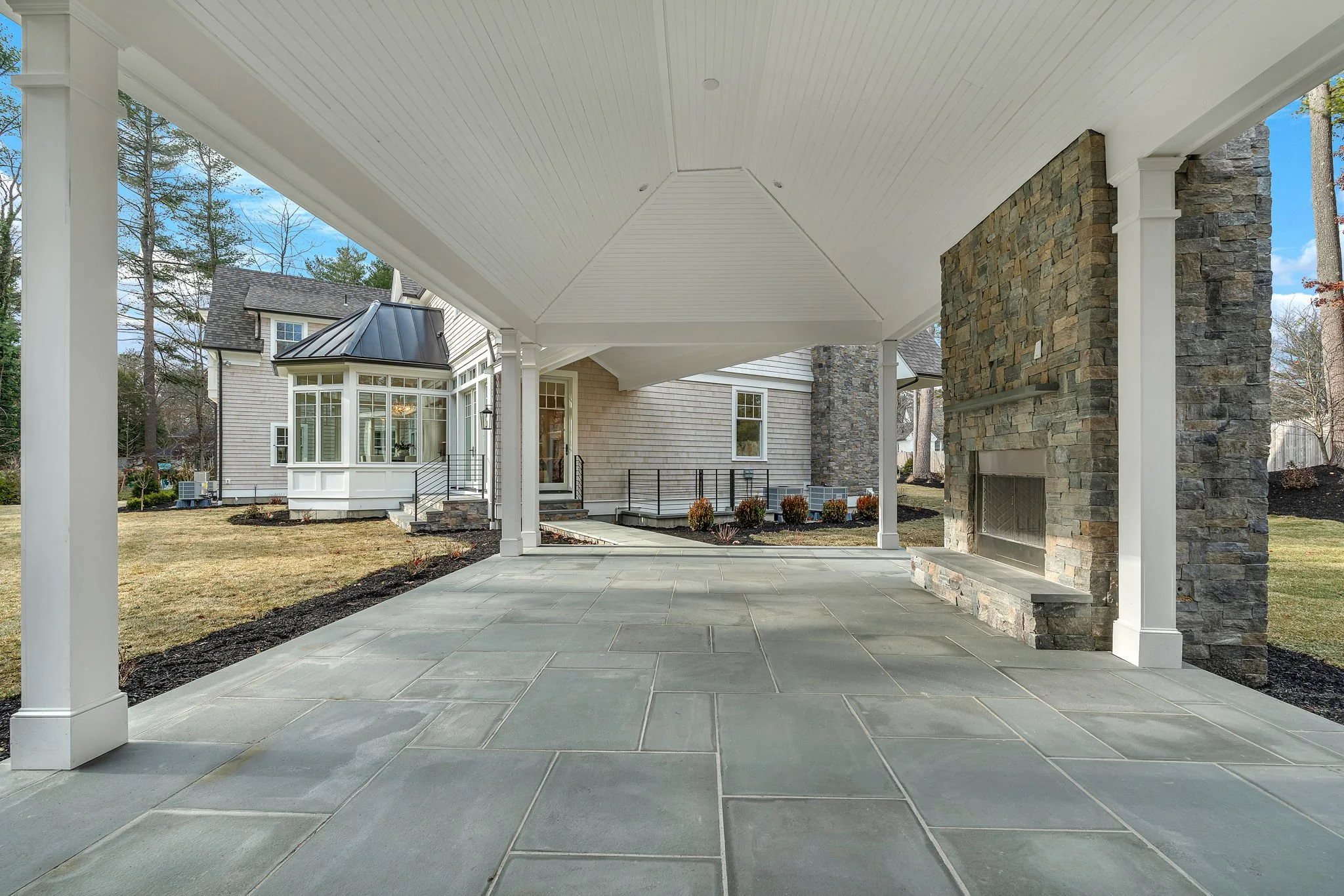Covered outdoor patio with stone fireplace, light-colored stone tiles, and a view of a house with a bay window and a lawn.