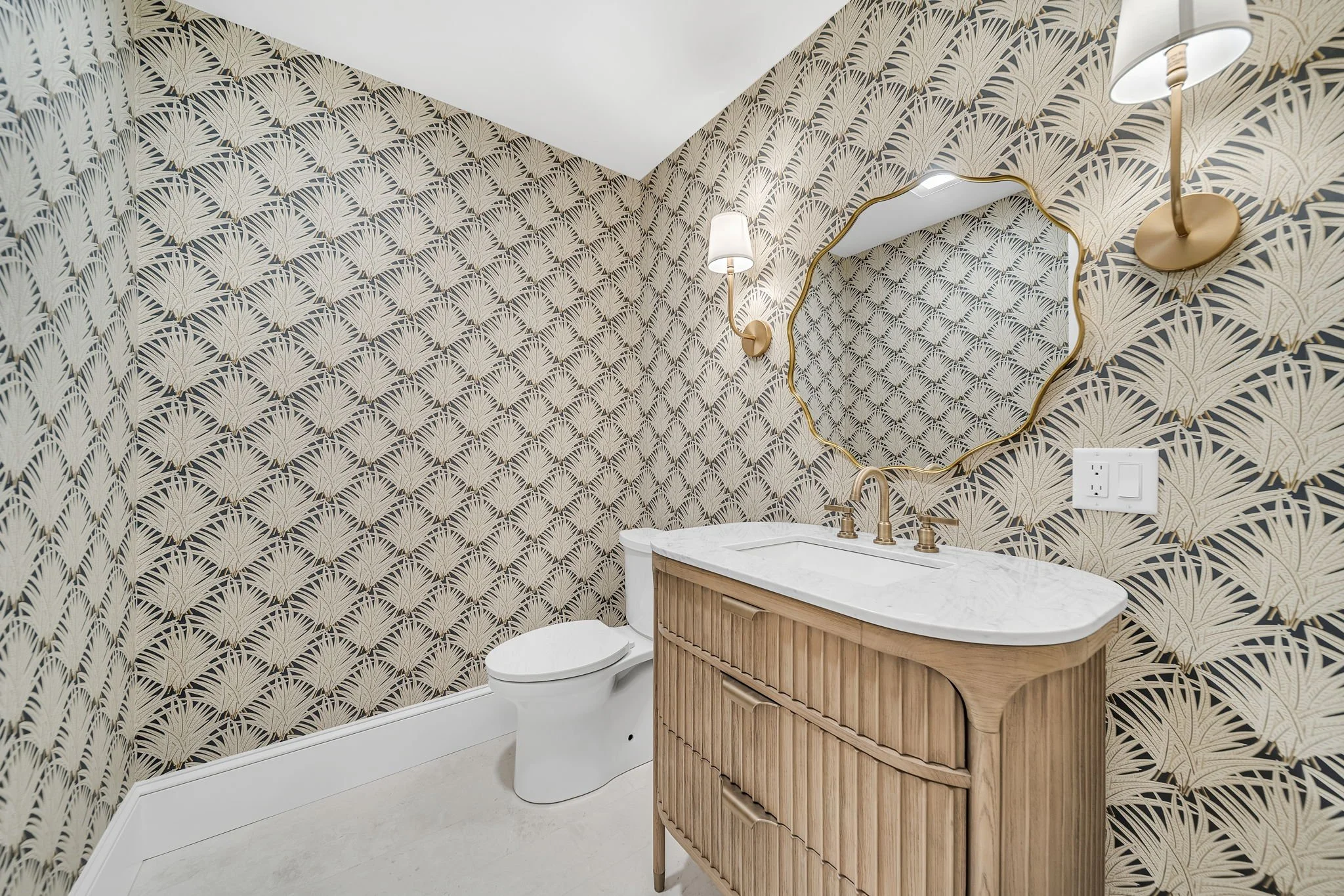 A small bathroom with beige and black patterned wallpaper, a white toilet, and a wooden vanity with a white marble top. There is a large irregularly shaped mirror above the vanity, and two gold wall-mounted lamps with white shades.