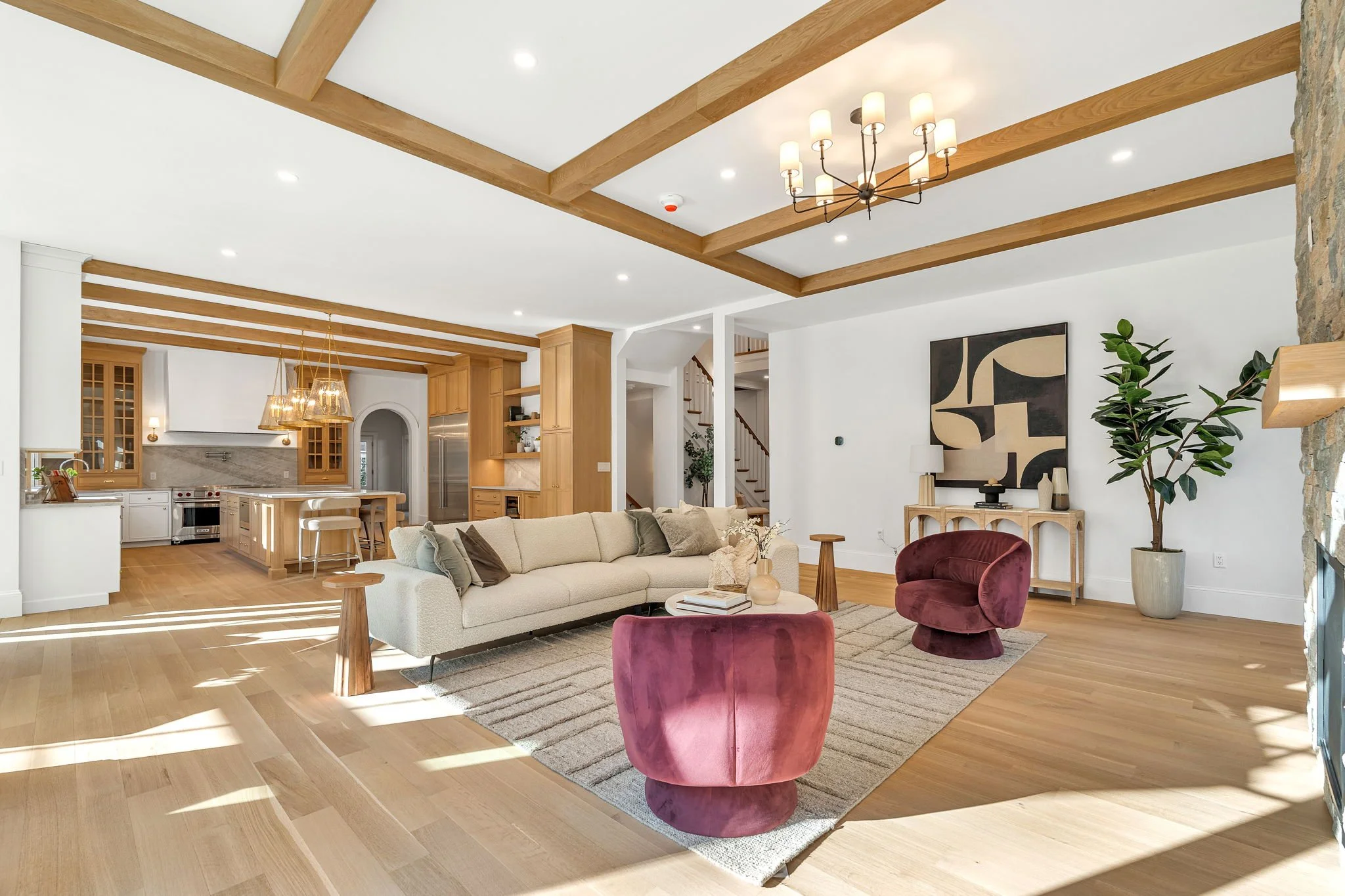 Living room with a beige sofa, two pink velvet chairs, a wooden coffee table, a rug, artwork on the wall, a potted plant, and a fireplace with stone details, in a bright, open space with wooden accents.
