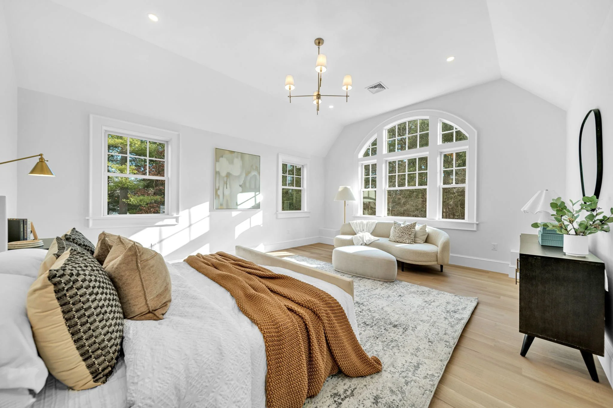 Bright and spacious bedroom with white walls and large windows, beige furniture, a beige sofa, a white bed with multiple pillows, a brown throw blanket, a black dresser, and wooden flooring.