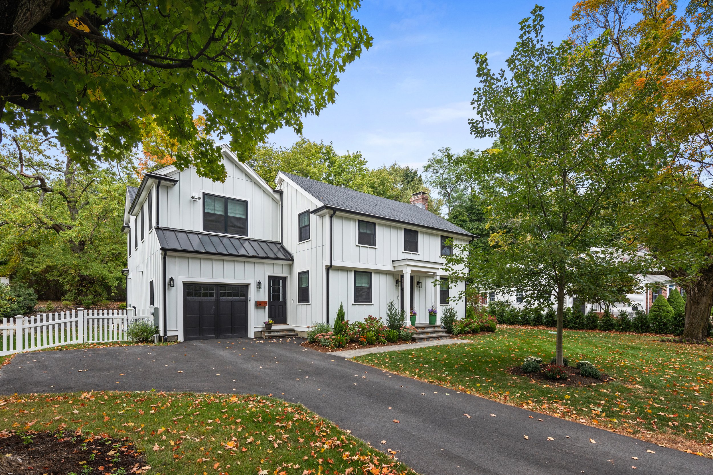 A modern white two-story house with black doors and windows, a black garage door, and a gray shingled roof, surrounded by a green lawn with trees and fallen leaves.