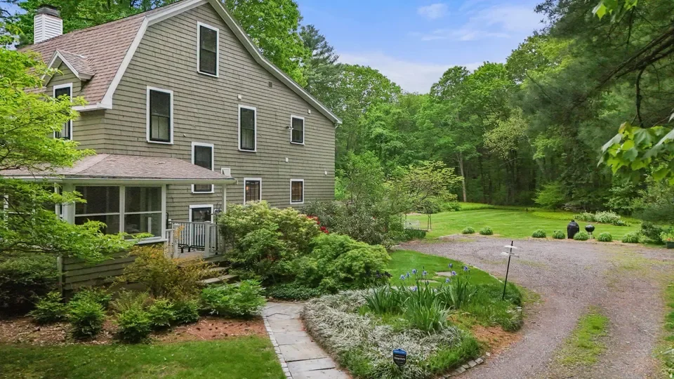 A large multi-story house with green siding, surrounded by lush greenery and a well-maintained garden. There is a paved pathway leading to the house, a gravel driveway, and a spacious yard with trees and bushes.