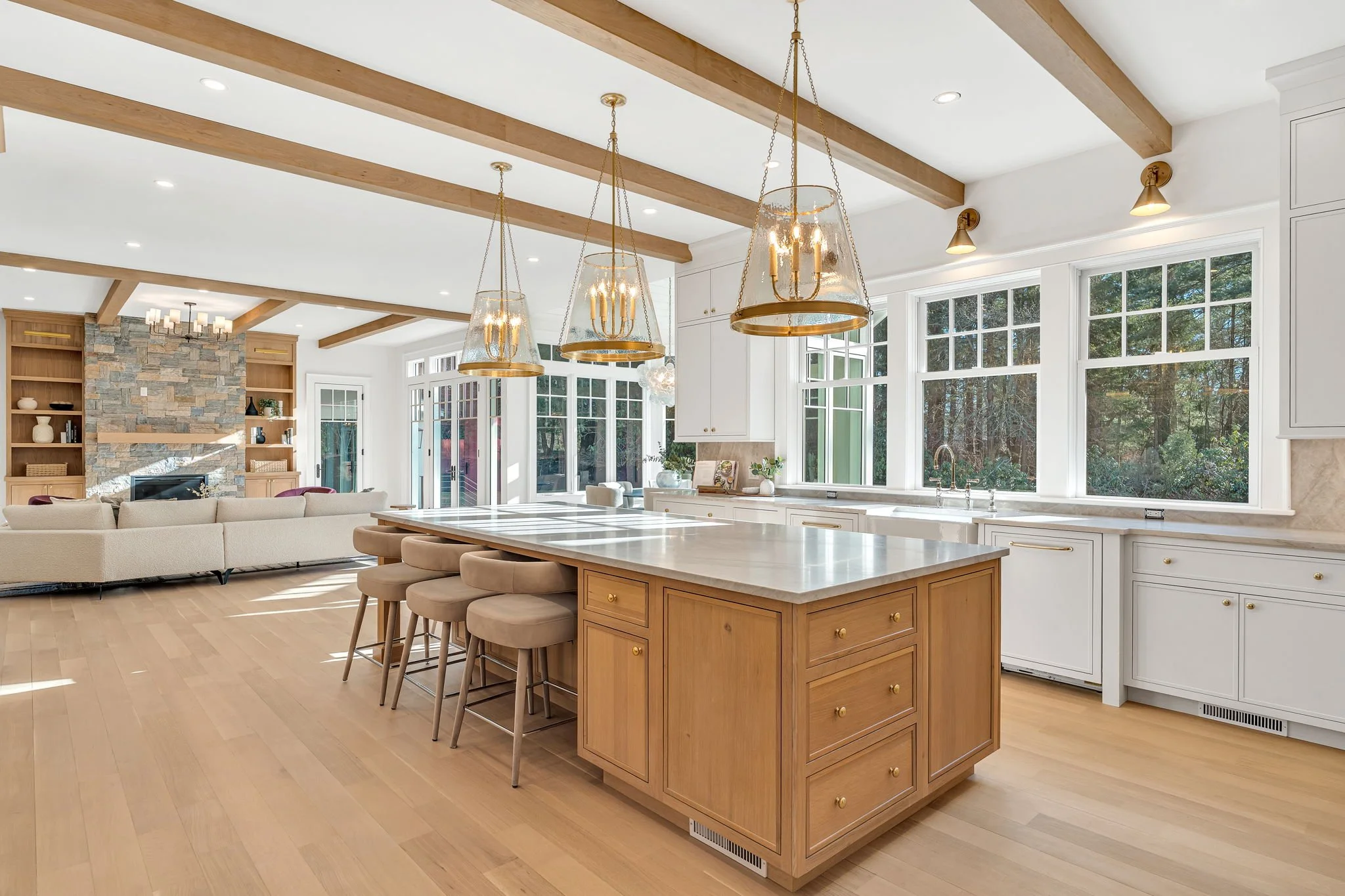 Bright open-concept kitchen and living room with large windows, wooden beams, island with barstools, and a stone fireplace.