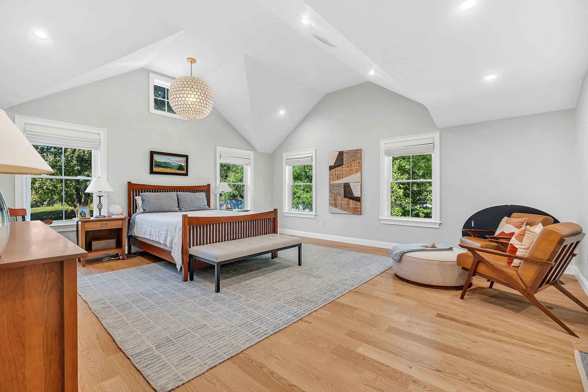 Bright bedroom with white walls and ceiling, wooden furniture, four windows with greenery outside, with a bed, bench at foot of bed, side tables, lamps, seated chairs, and wall art.