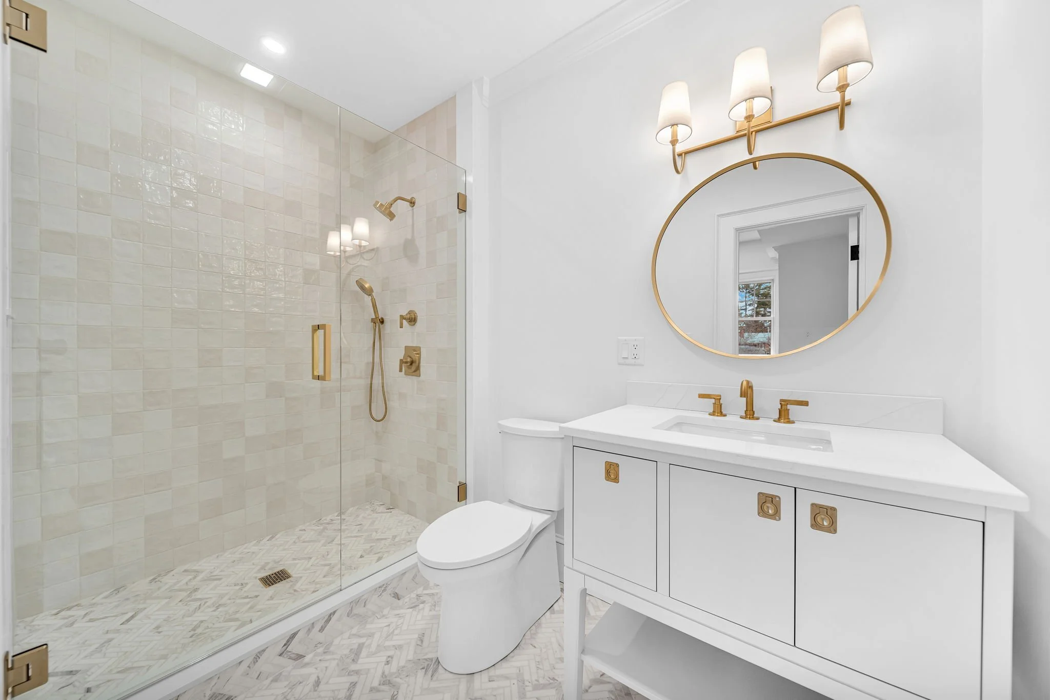 Modern white bathroom with a glass-enclosed shower with gold fixtures, a white vanity with gold hardware, a round mirror, and a three-light wall sconce.