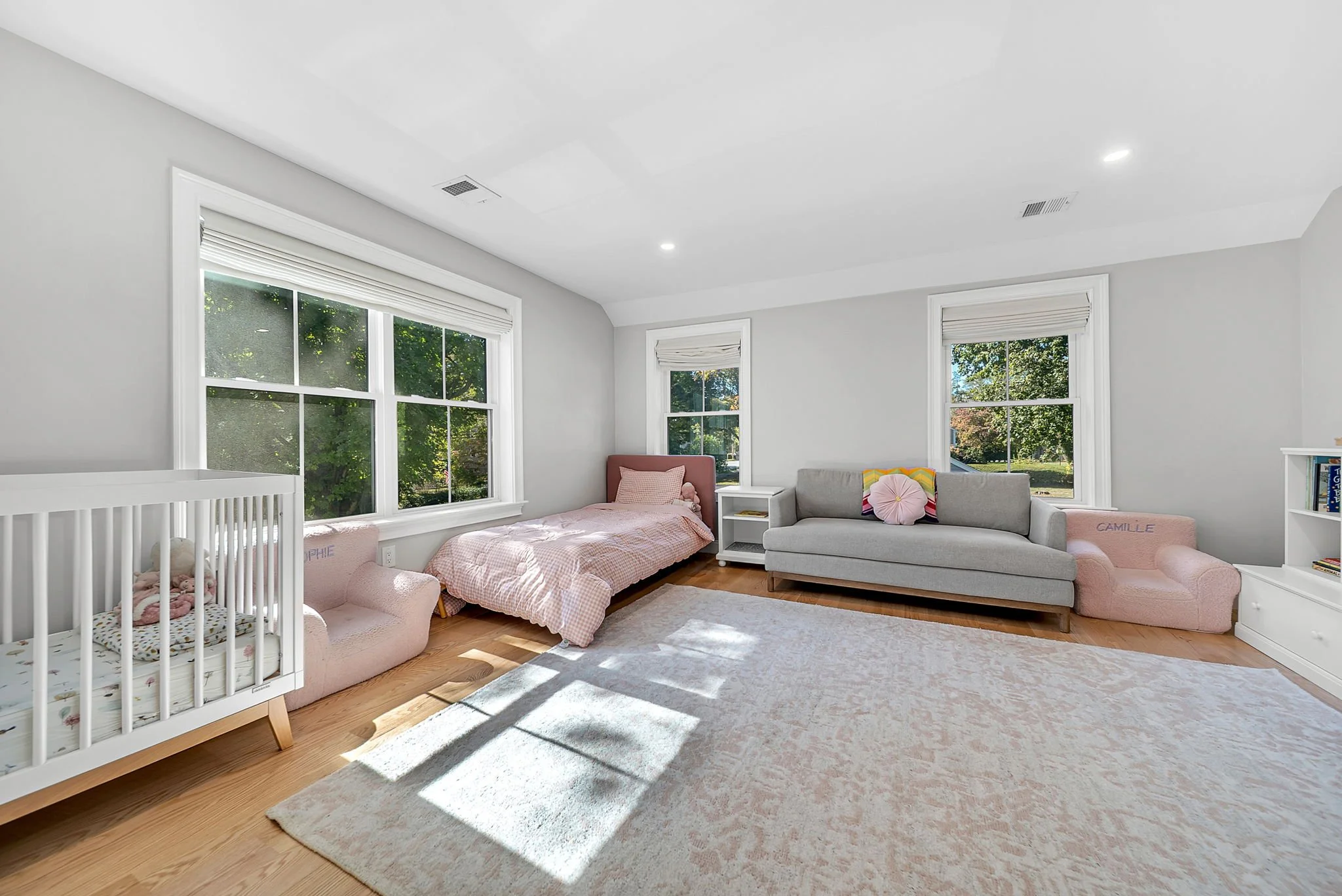 Bright nursery room with large windows, white walls, hardwood floors, a crib, a twin bed with pink bedding, a gray sofa, and pink armchairs.