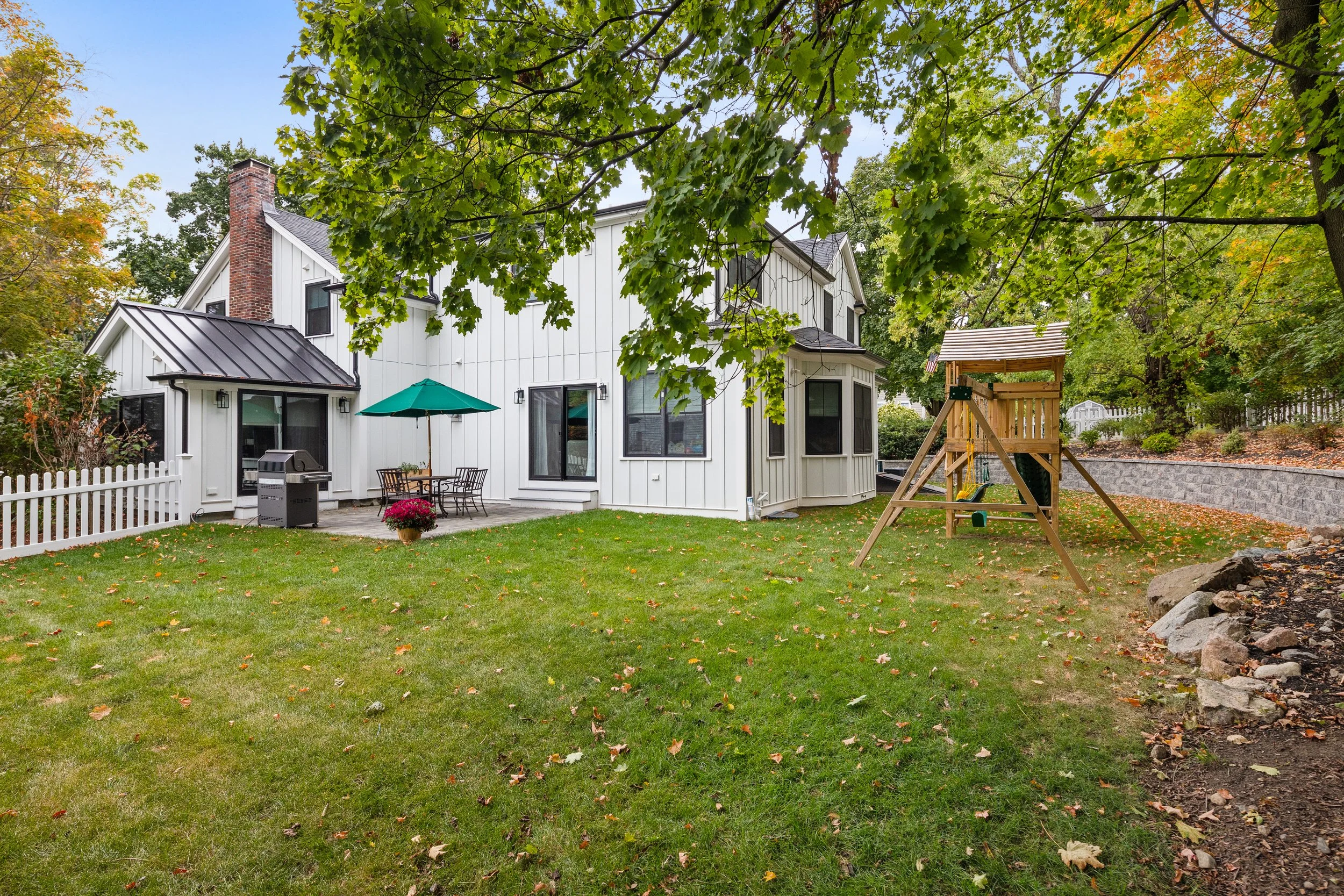 Backyard of a white house with a green lawn, trees with green leaves, outdoor seating with an umbrella, a grill, and a wooden playhouse with a slide.
