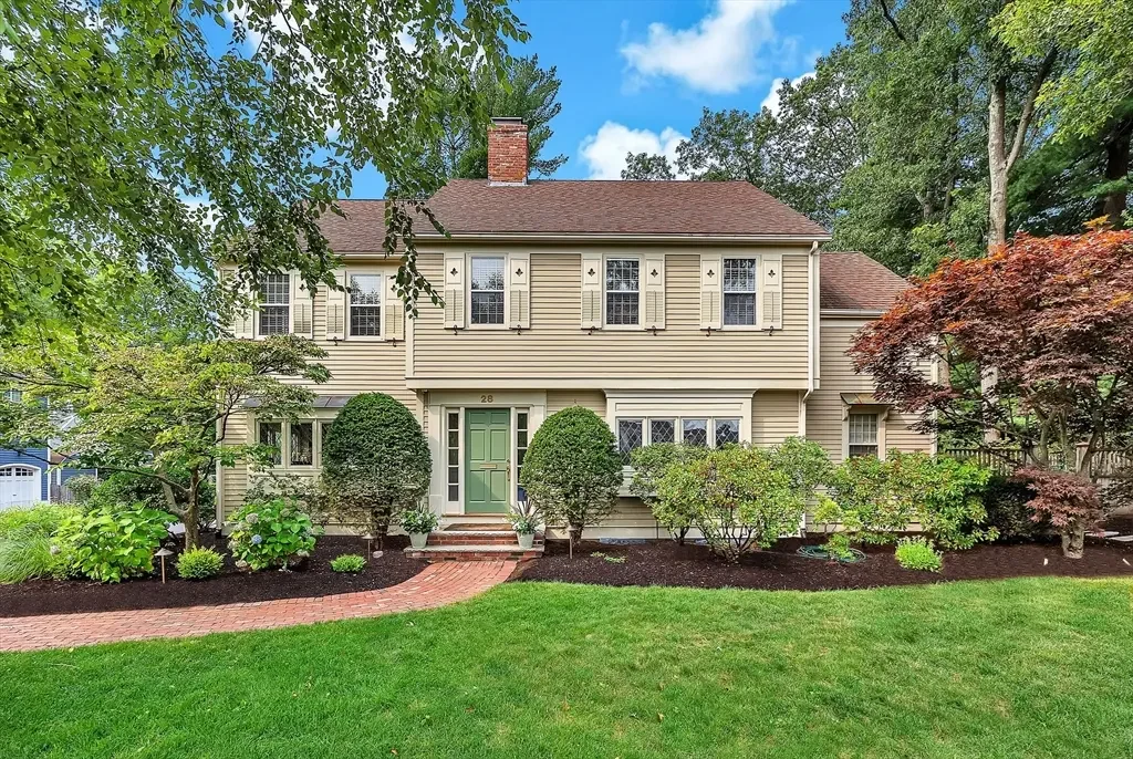 Front view of a two-story house with beige siding, green front door, and white window shutters, surrounded by a landscaped yard with green grass, bushes, and trees.