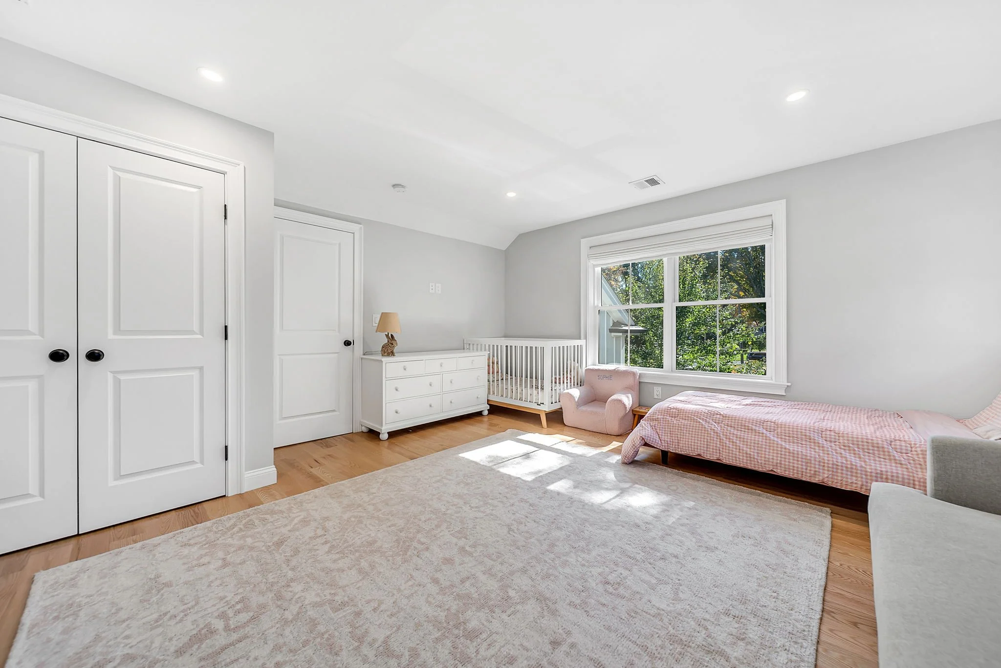 Bright nursery with white walls, large window with greenery outside, a pink bed with gingham blanket, a small pink armchair, a white dresser with a lamp, and a bassinet near the window.