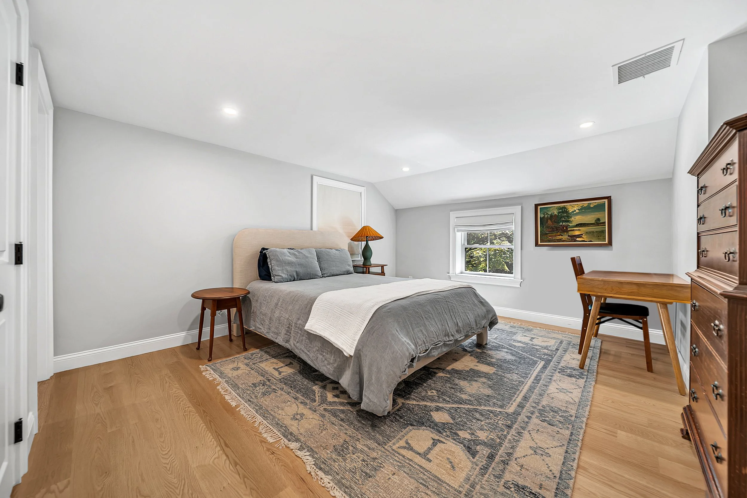 A bedroom with a bed, nightstand, lamp, desk, and painting, featuring white walls, hardwood floors, and a window with greenery outside.