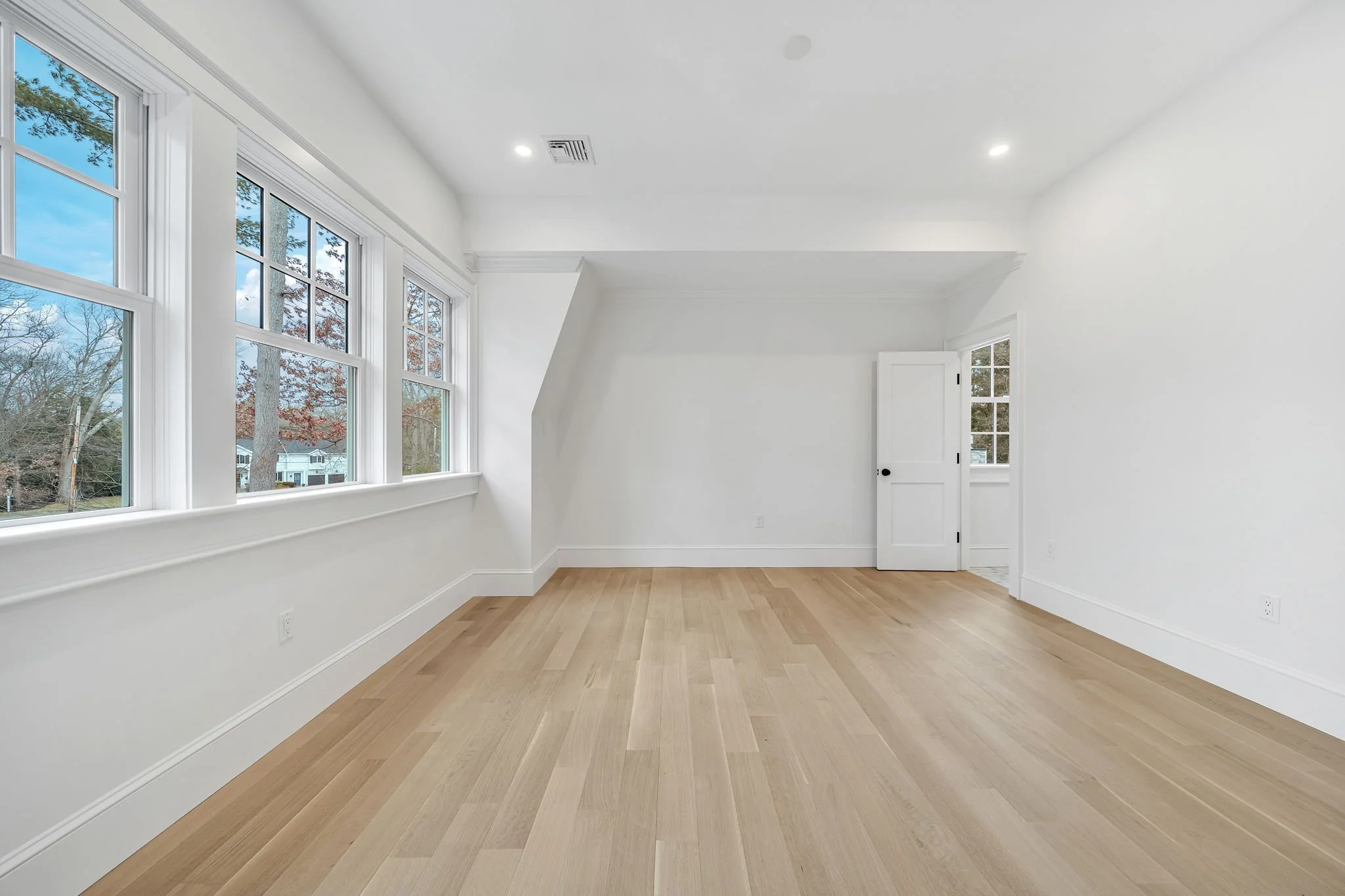 Empty room with white walls, light wood flooring, multiple large windows, a white door, and ceiling lights.