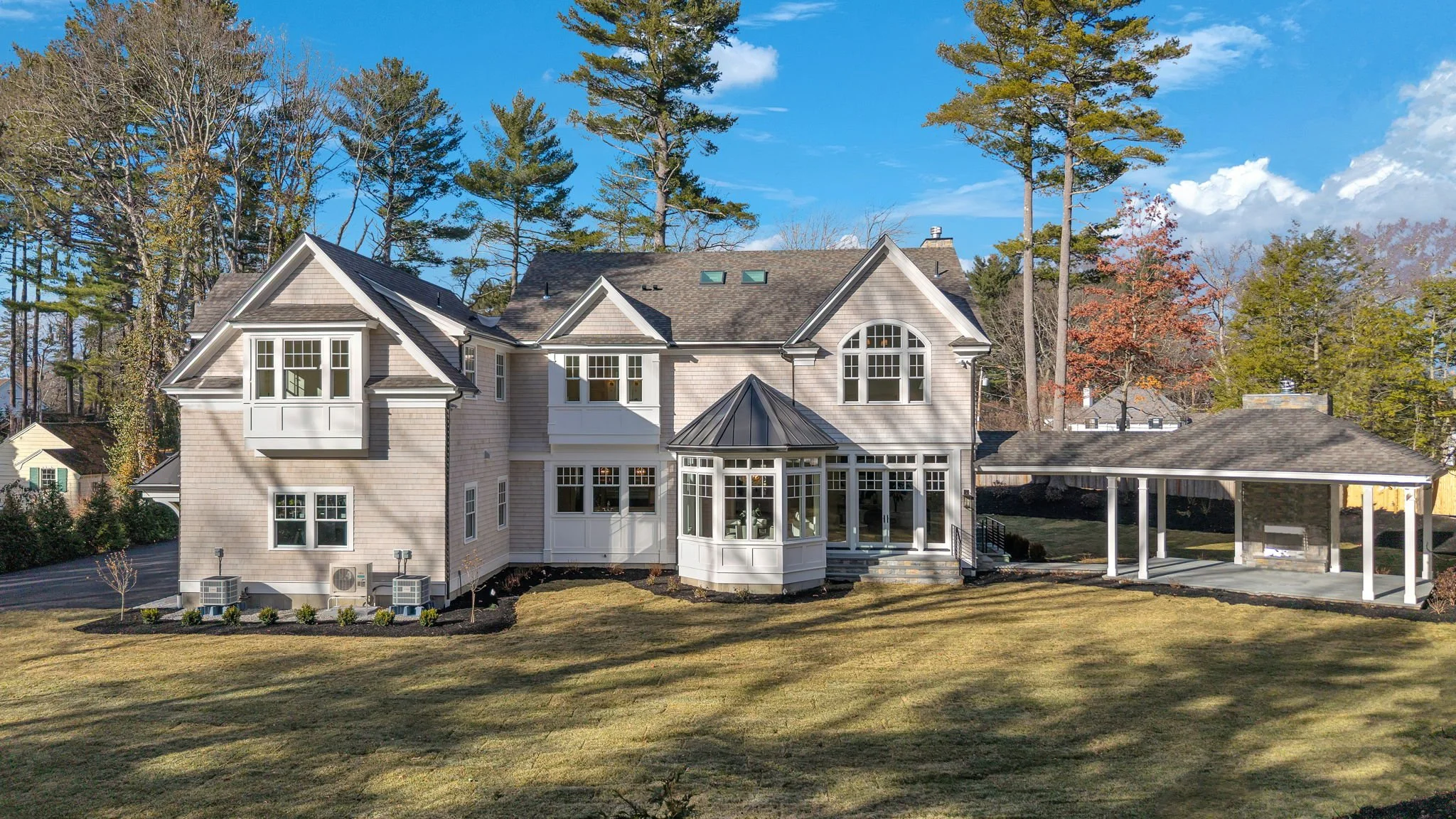 Large, modern two-story house with beige siding and multiple windows, including a glass-enclosed porch, surrounded by trees and a well-maintained yard.