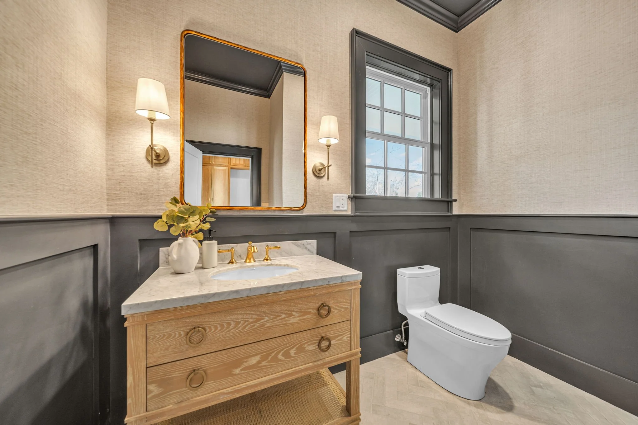 A modern bathroom with a wooden vanity, marble countertop, gold fixtures, a rectangular mirror, wall sconces, a window with dark trim, and a white toilet.