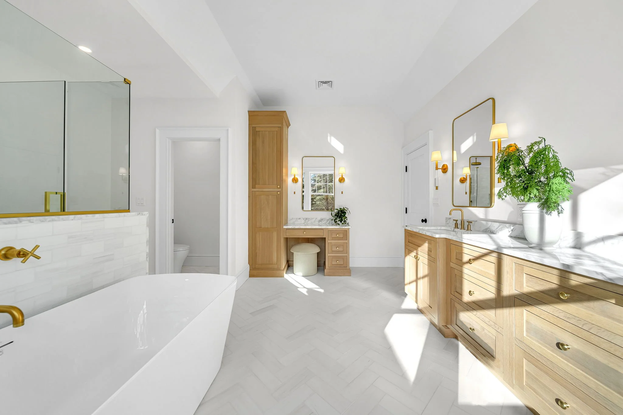 Bright bathroom with a large freestanding bathtub, wooden cabinets, a vanity with a marble countertop, and a small makeup area with a mirror and stool. Features include white walls, gold fixtures, a large mirror, wall-mounted lights, and a shower wit