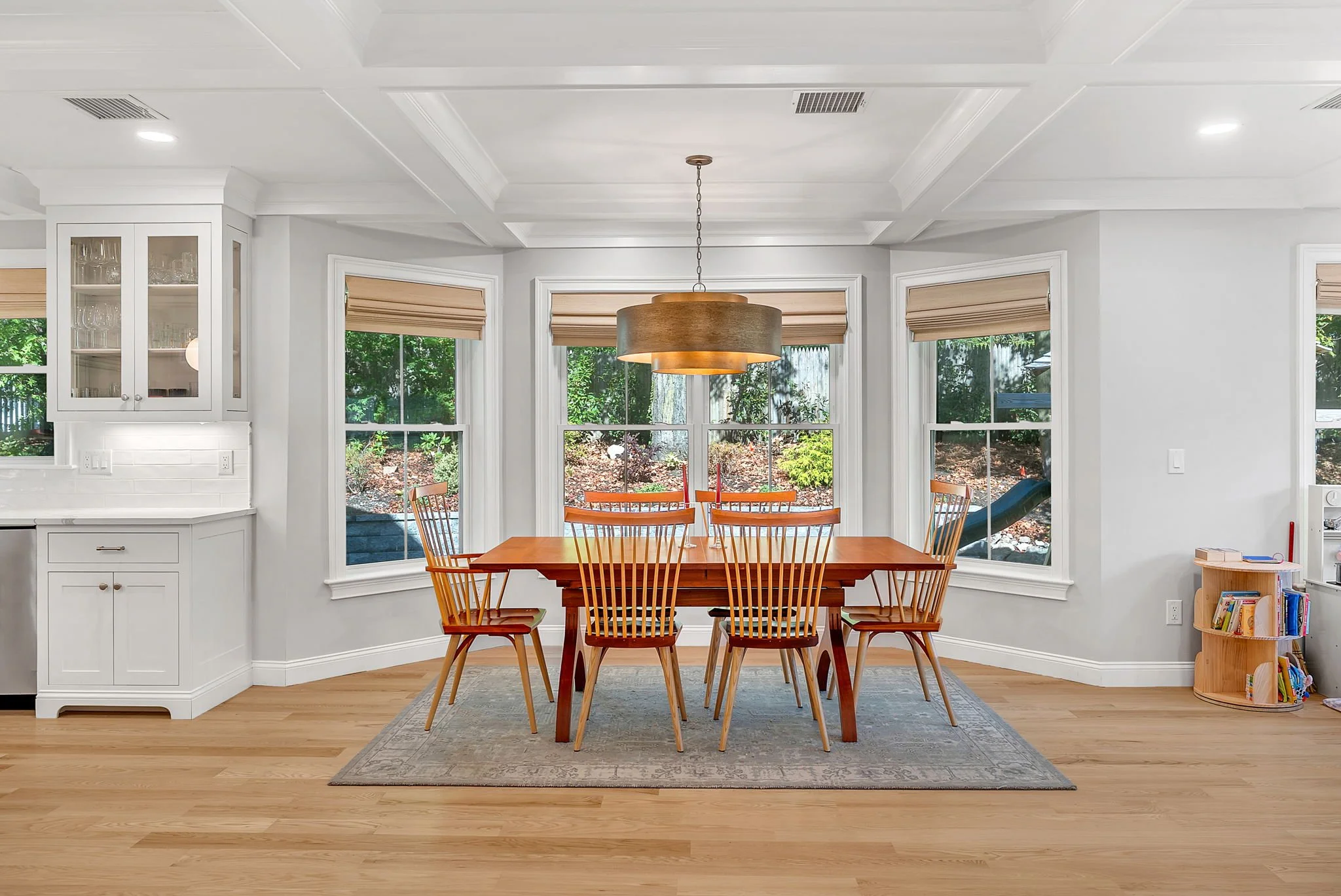 Bright dining room with a wooden table, six chairs, a modern hanging light fixture, three large windows with beige shades, a white kitchen corner with cabinets and glassware, a small book shelf, and a view of trees and a slide outside.