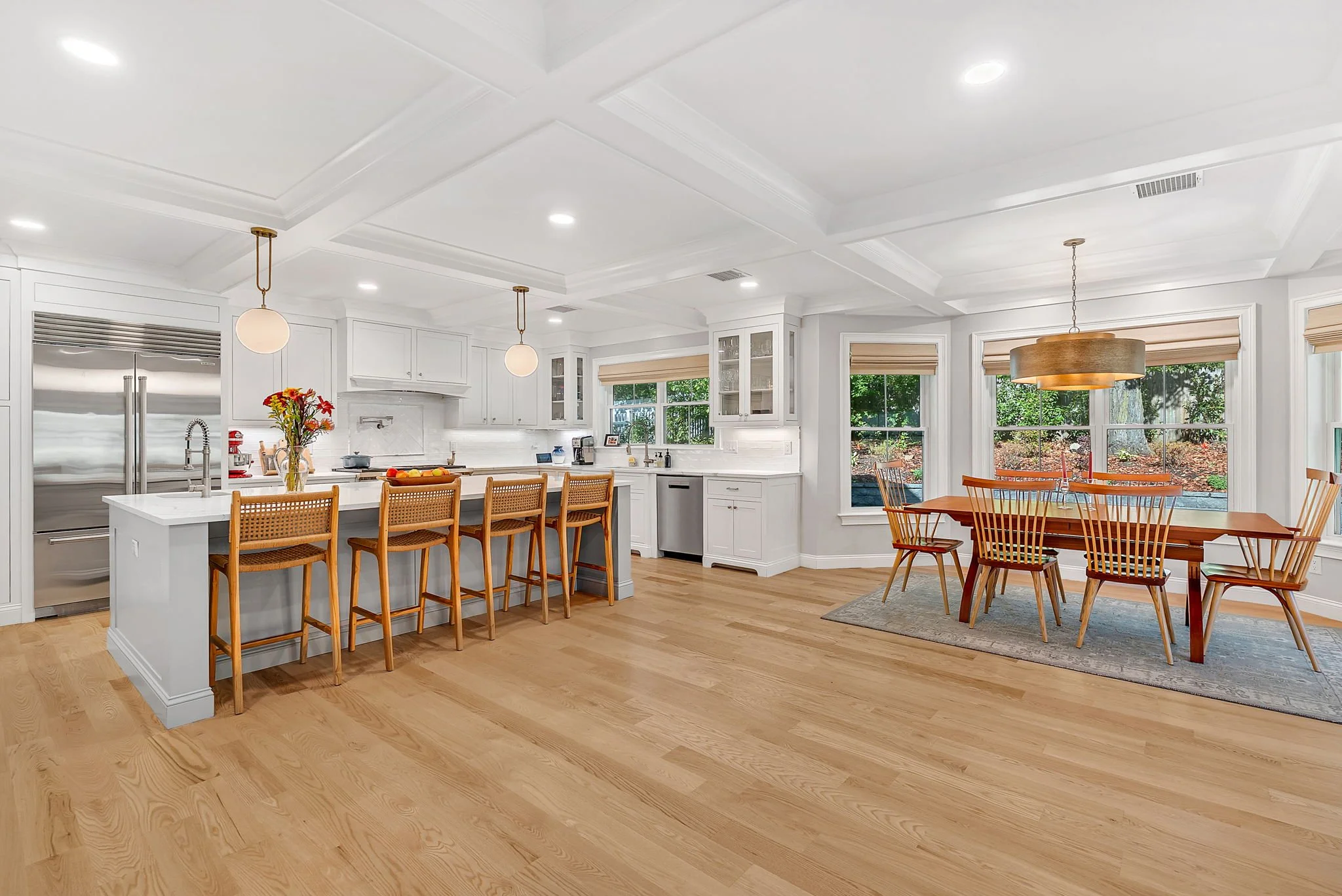 Bright open kitchen and dining area with white cabinets, a kitchen island with five wooden bar stools, large windows with blinds, and a wooden dining table with six chairs, a hanging light fixture, and a view of greenery outside.