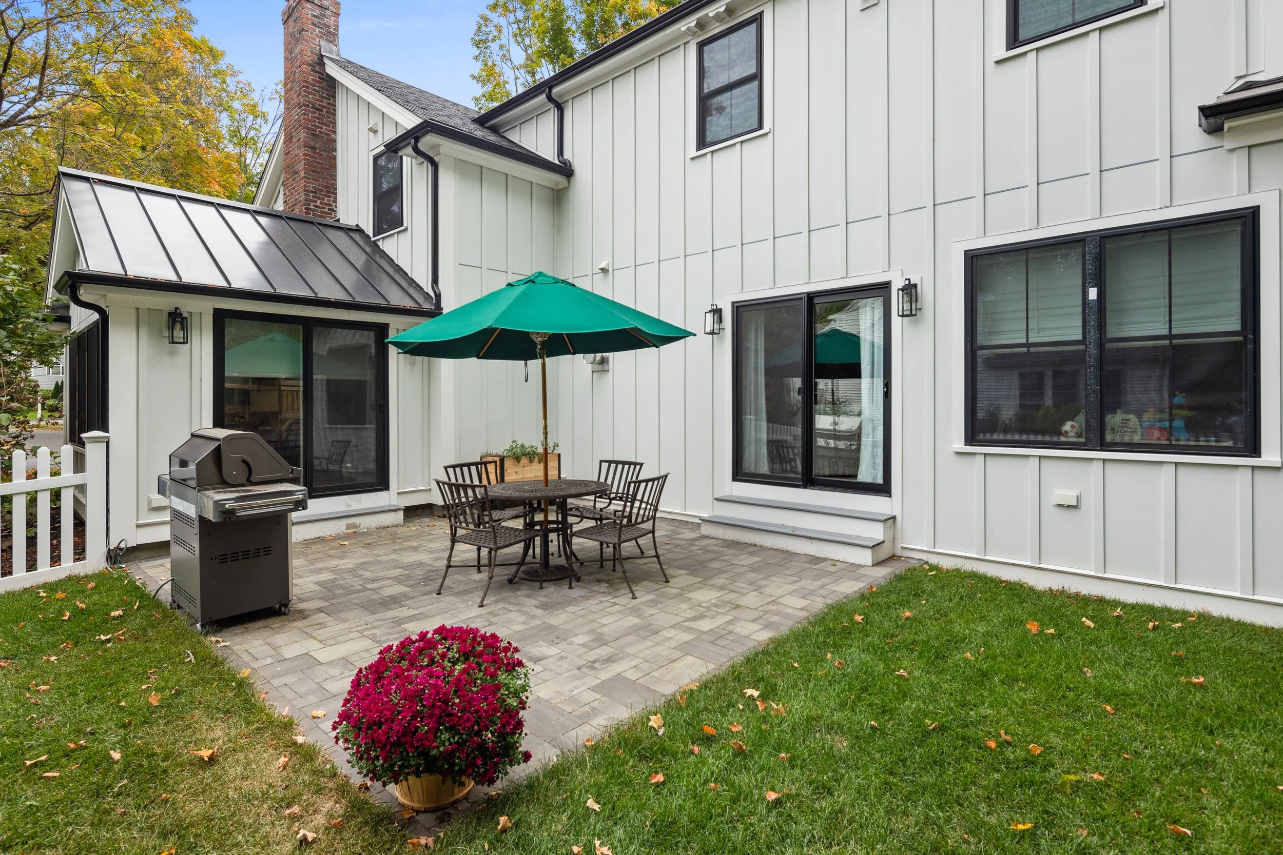 Backyard patio area with a round table and four chairs with a large green umbrella, a charcoal grill, a flowering plant in a pot, grass, and large windows on a white house with black framing.