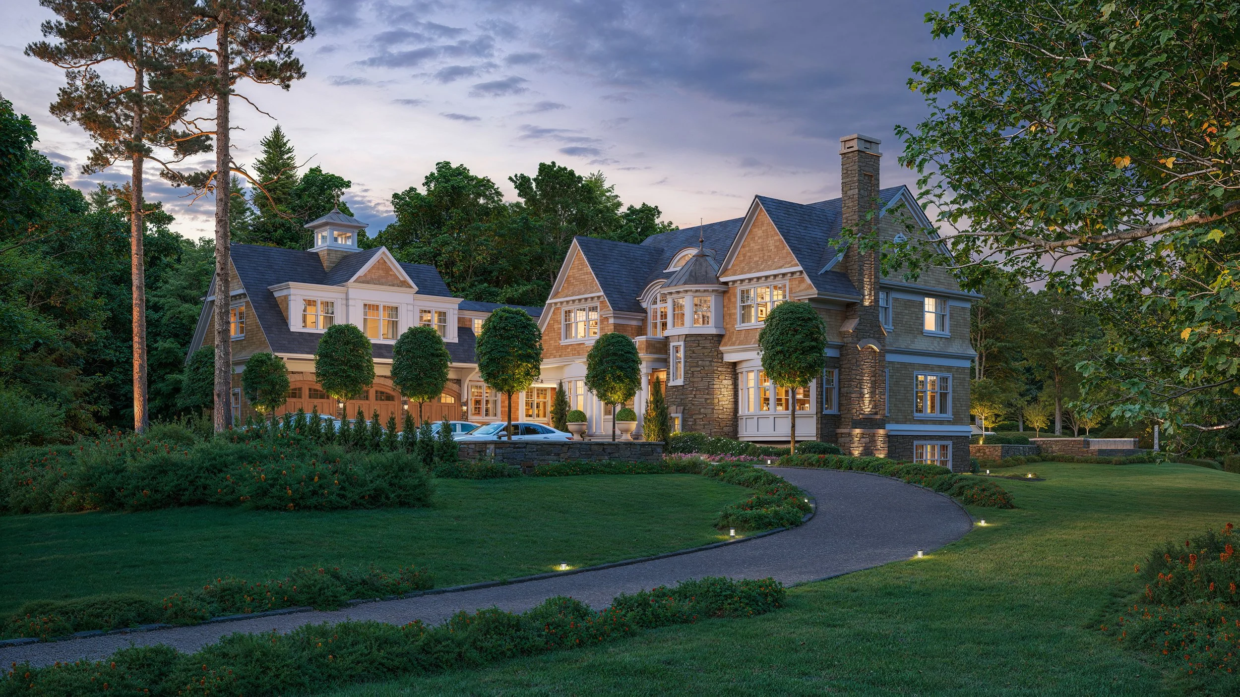 Large, elegant house with stone and siding exterior, multiple gables, turret, and large windows, surrounded by landscaped lawn and trees at dusk.