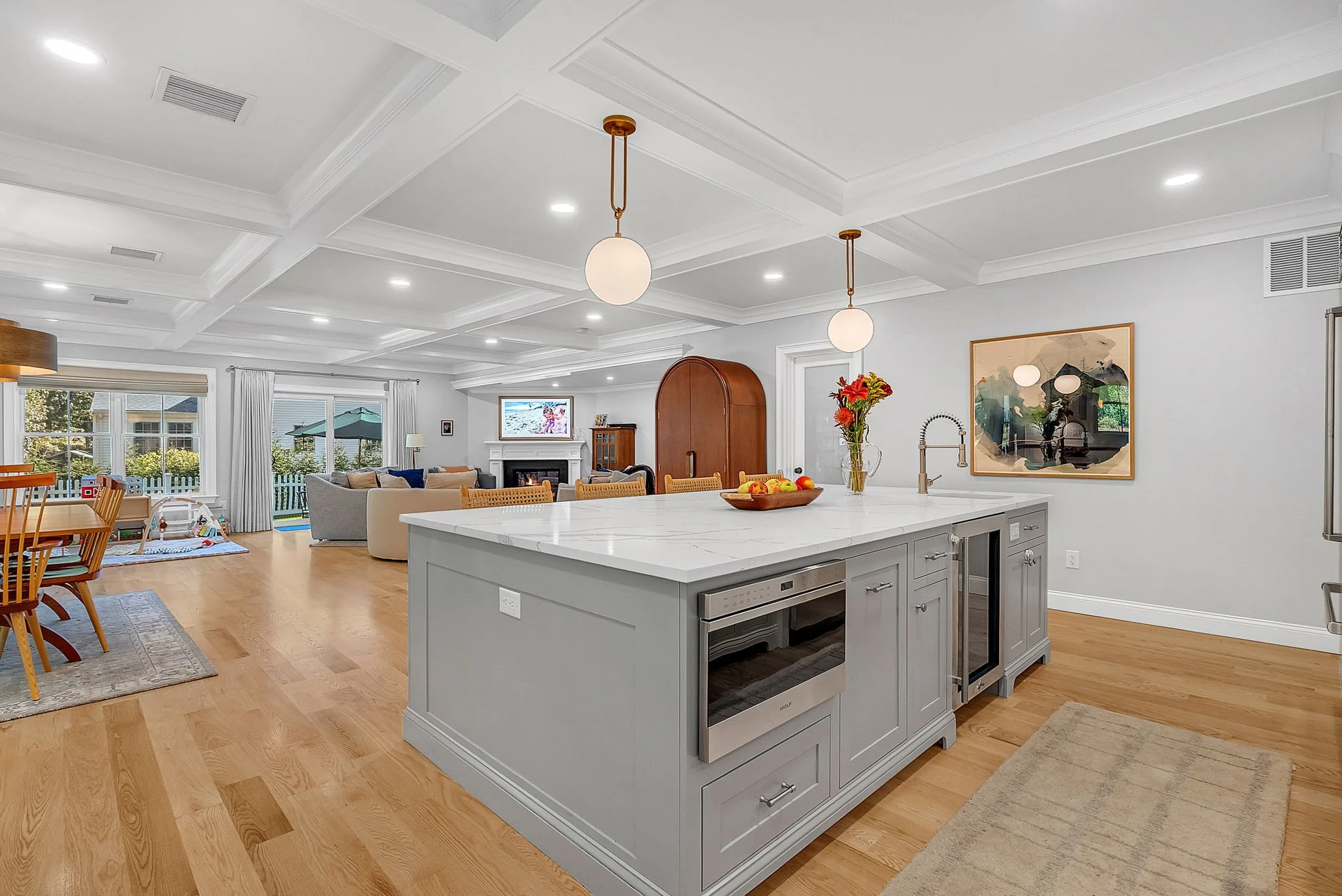Open concept living room and kitchen with white walls, wooden flooring, and modern furnishings. Kitchen island with a white marble top, pendant lights, and a fruit bowl. Living area with sofas, fireplace, and windows with curtains.