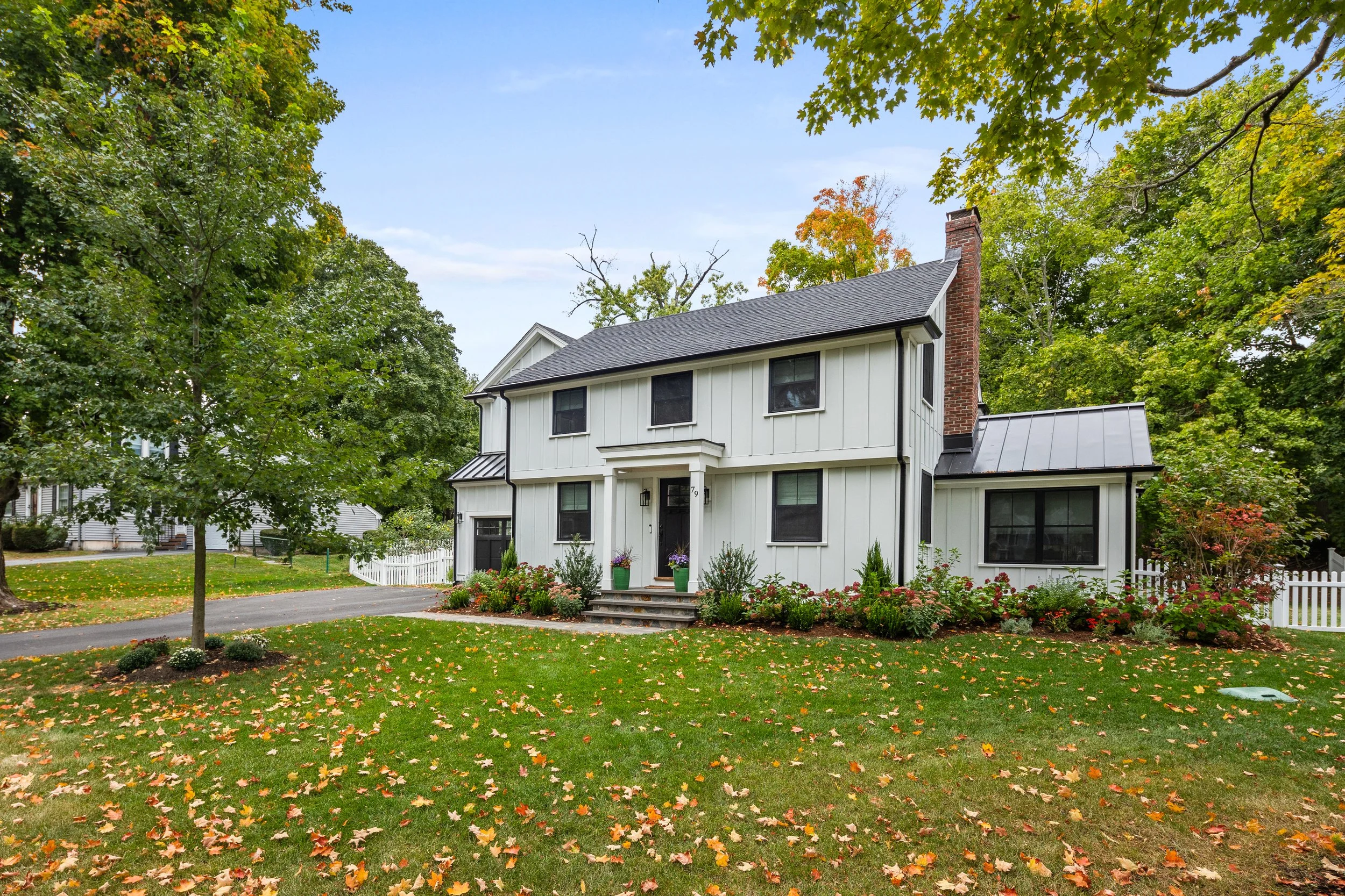 A two-story white house with black window frames and a brick chimney, surrounded by a well-maintained lawn with colorful fallen leaves, trees with green and orange foliage, and a small garden with flowers in front.