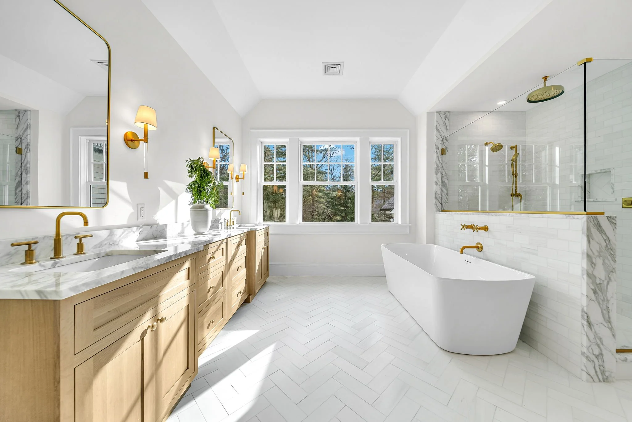 Bright modern bathroom with double vanity, large mirror, gold fixtures, free-standing bathtub, and a walk-in shower with gold fixtures and marble accents.