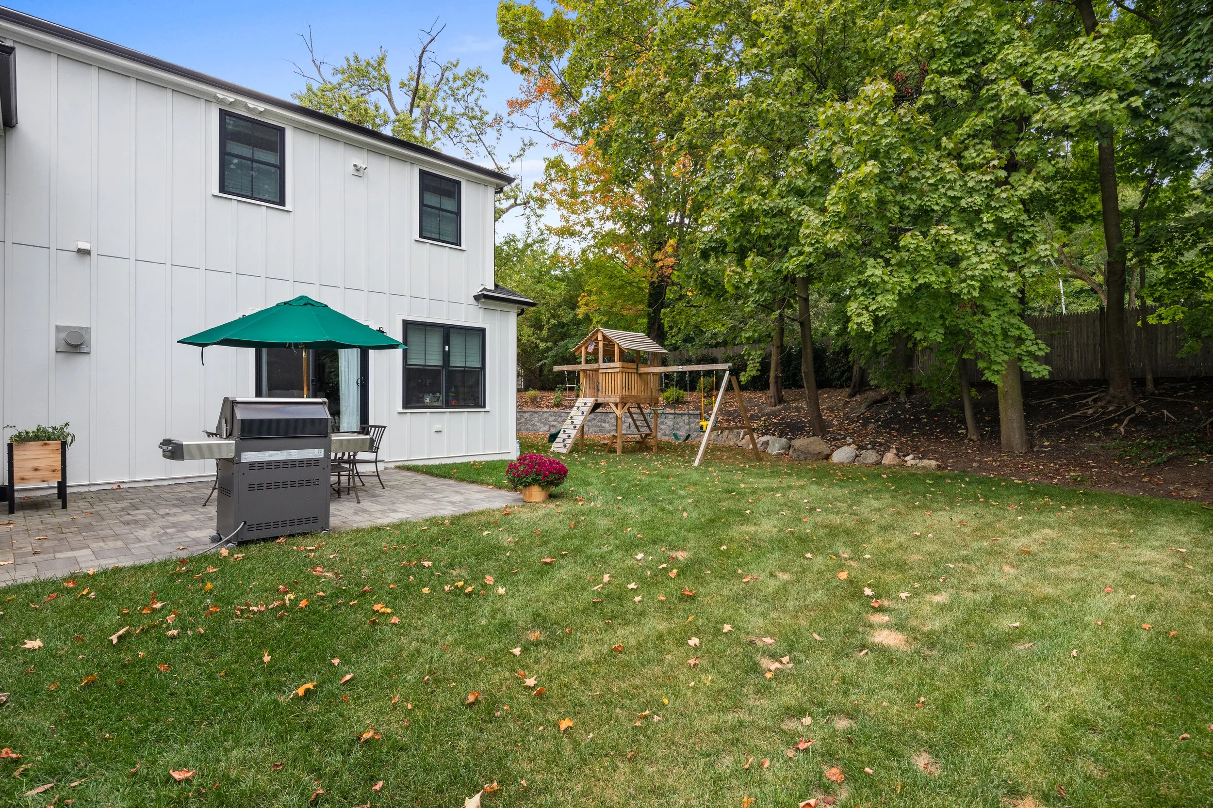 Backyard with a white house, a paved patio with a grill and patio furniture under a green umbrella, a small wooden playset with a slide and swings, a potted plant with pink flowers, lush green grass with scattered leaves, and a line of trees and a wooden fence in the background.