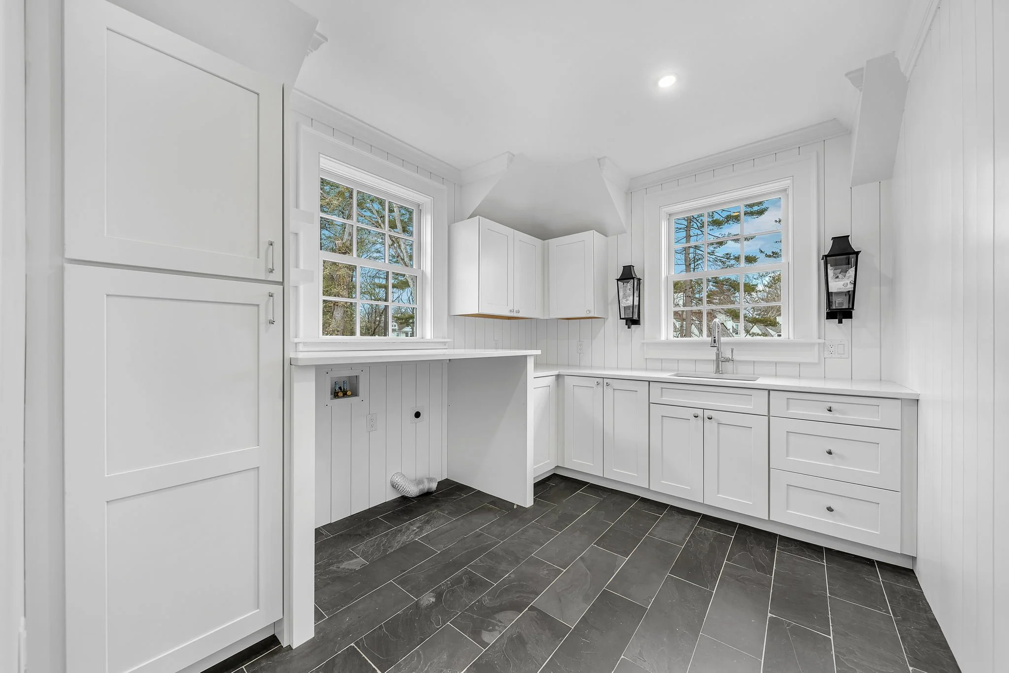 White kitchen with two large windows, white cabinets, black floor tiles, and black wall lanterns.
